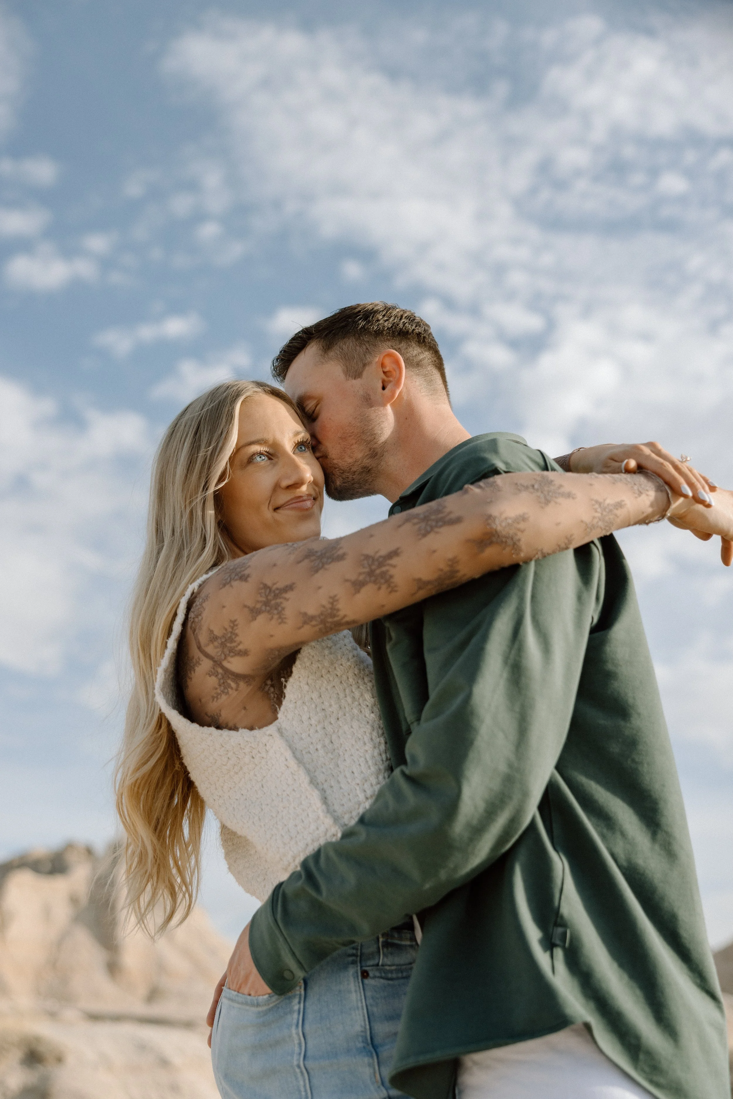A woman and man embrace outdoors against a blue sky with scattered clouds, with the woman looking at the man as he kisses her forehead.