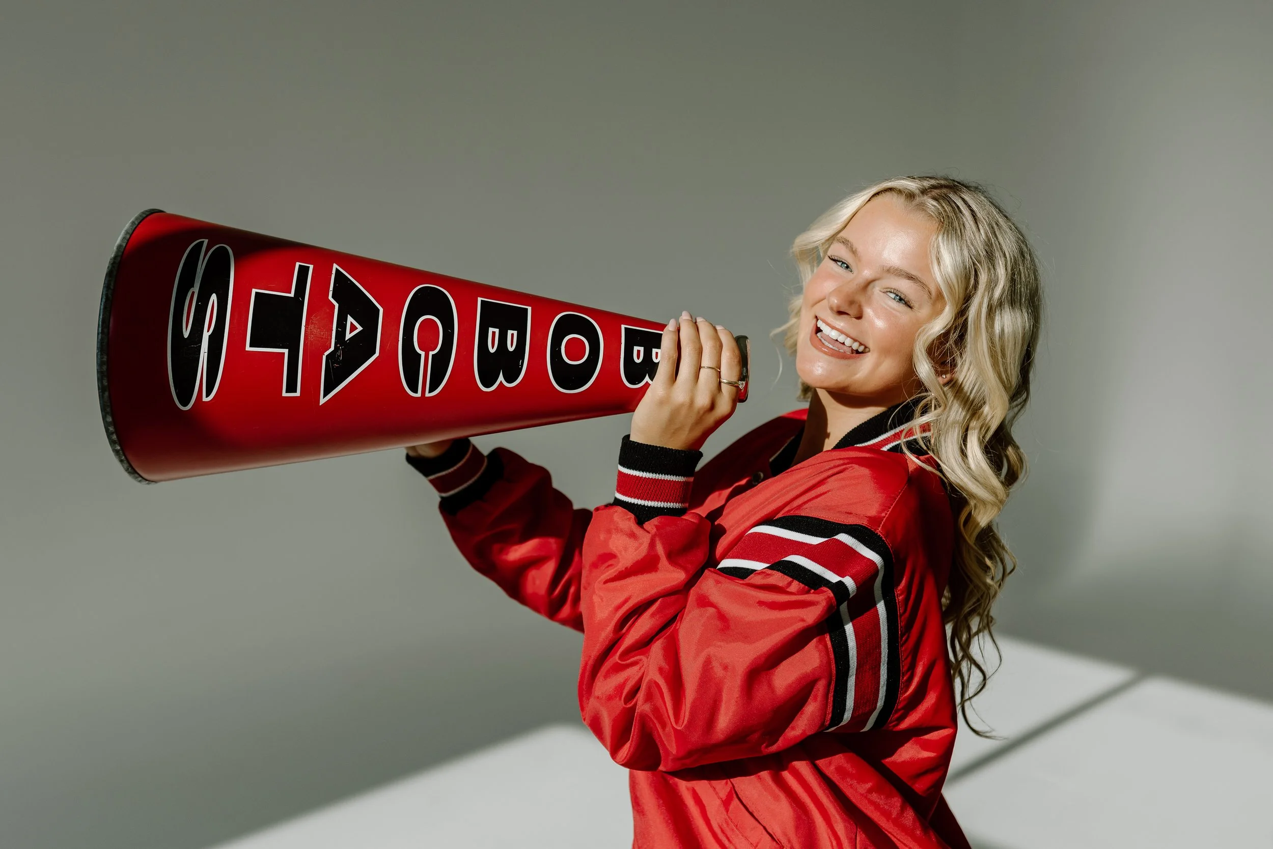 A woman with blonde curly hair smiling and winking while holding a red thunderstick with the words 'GO BUCKS' printed on it, wearing a red and black sports jacket.