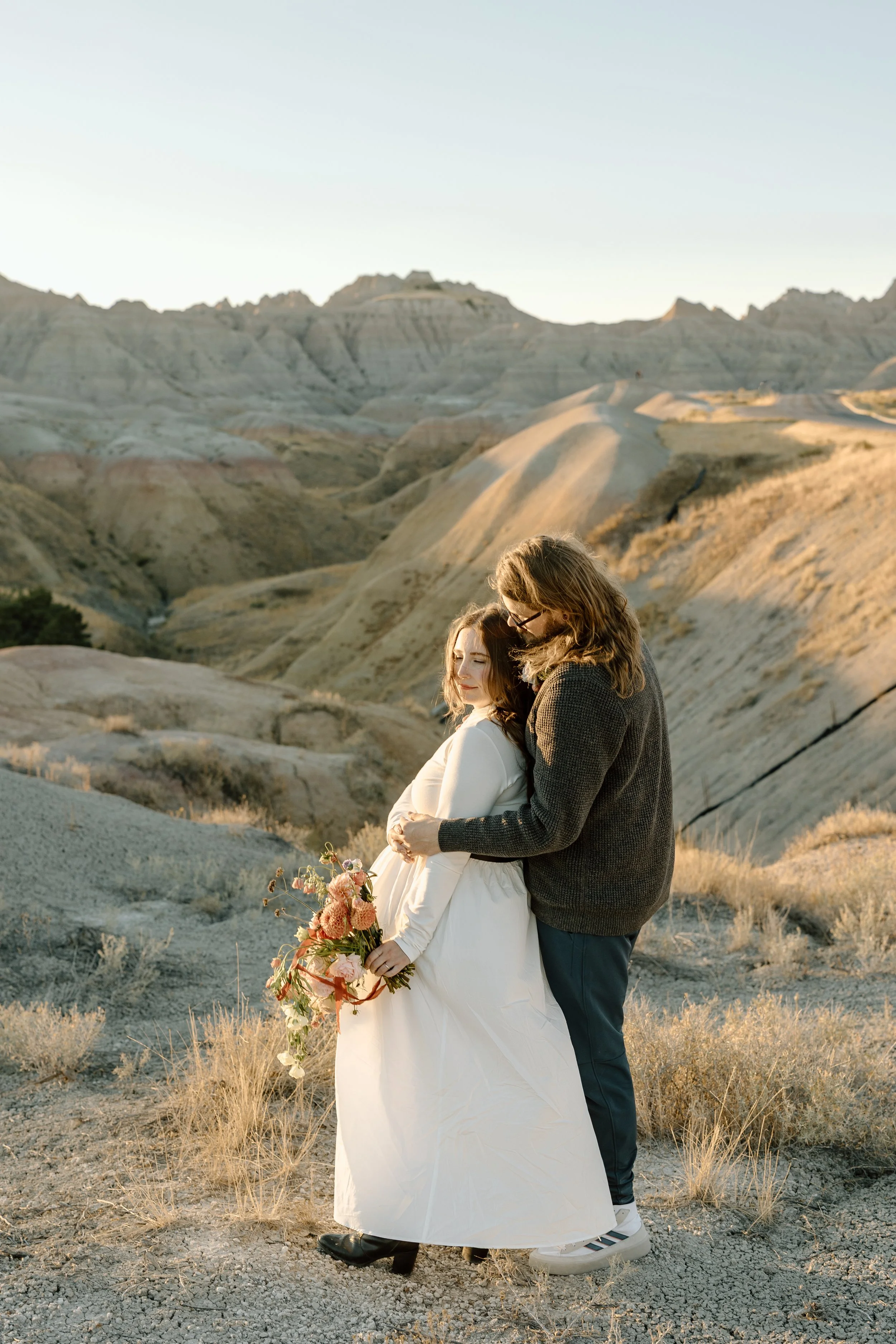 A couple embracing in a desert landscape with mountains in the background, the woman holding a bouquet of flowers.