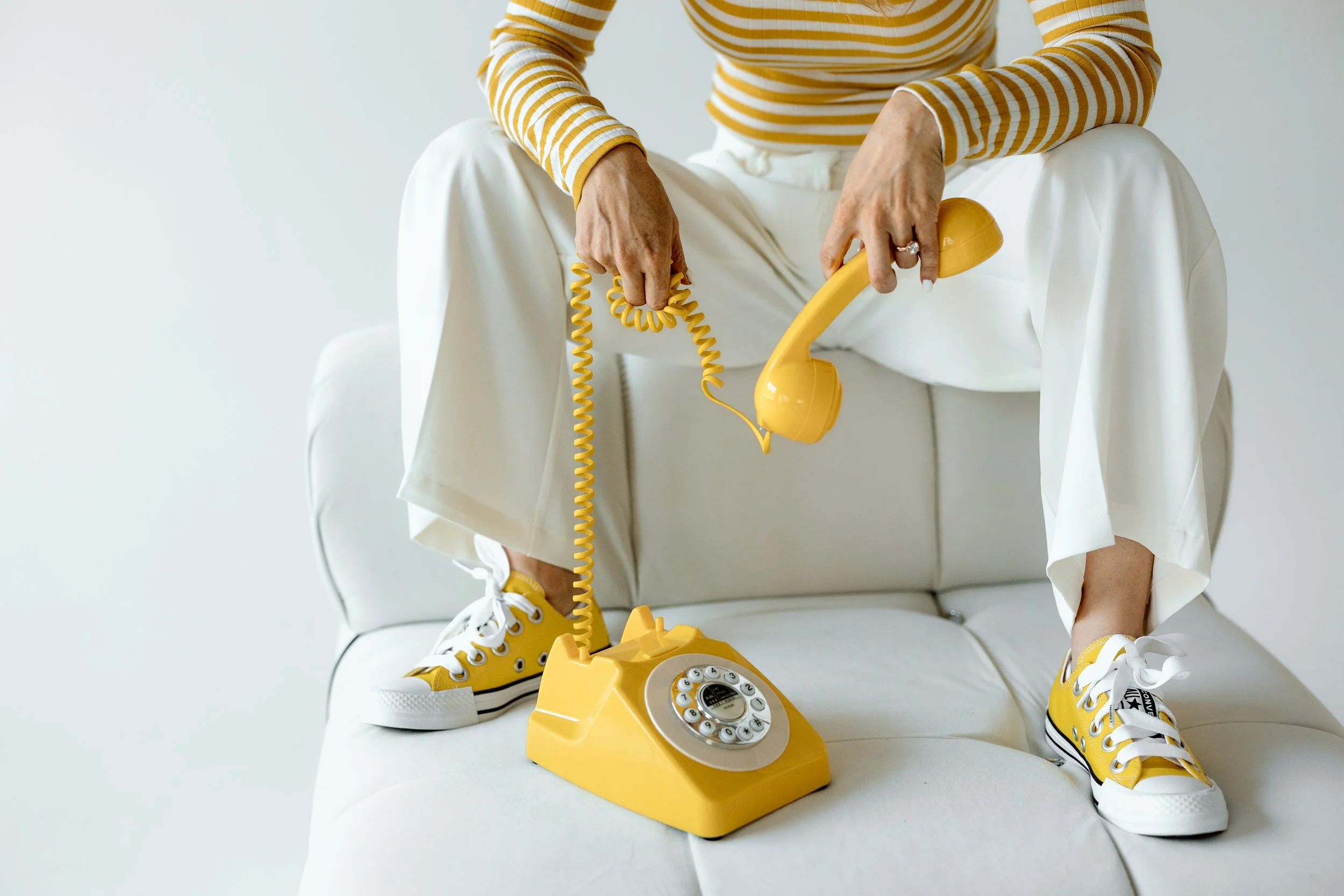 Person in yellow striped shirt and white pants sitting on white sofa, holding a yellow retro rotary phone with the receiver in one hand and the cord in the other.