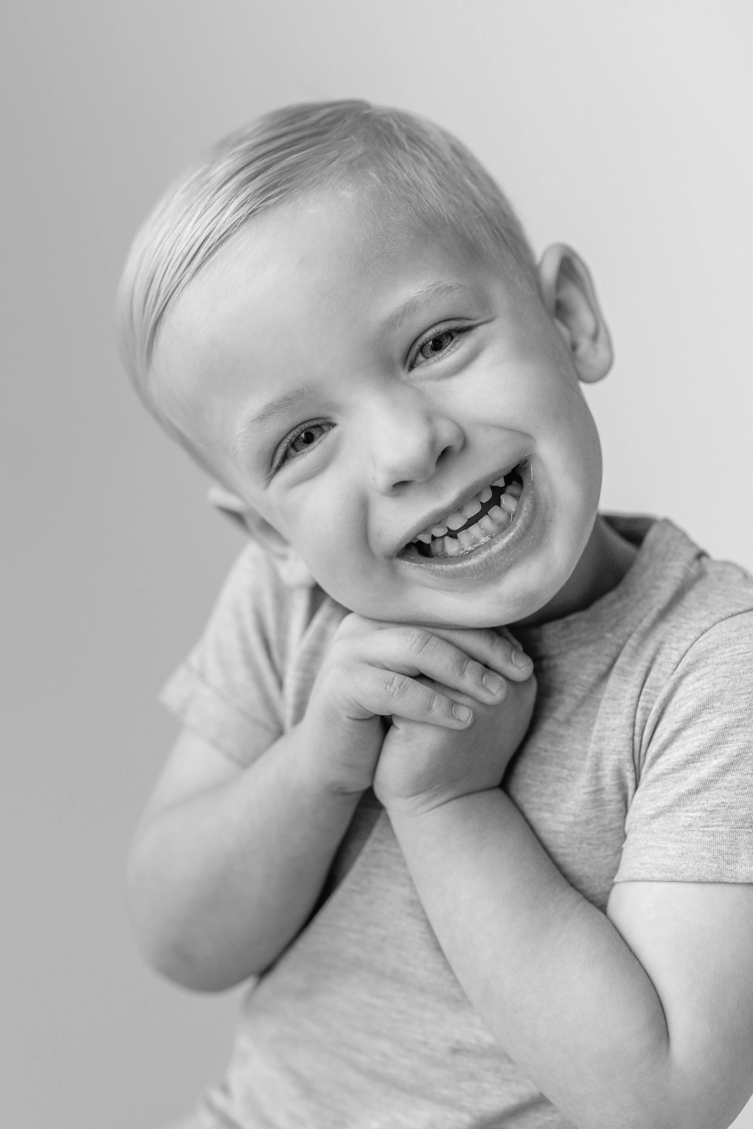 Black and white photo of a happy young boy with short, swept hair, smiling with his teeth showing, holding his chin with both hands, wearing a short-sleeved shirt.