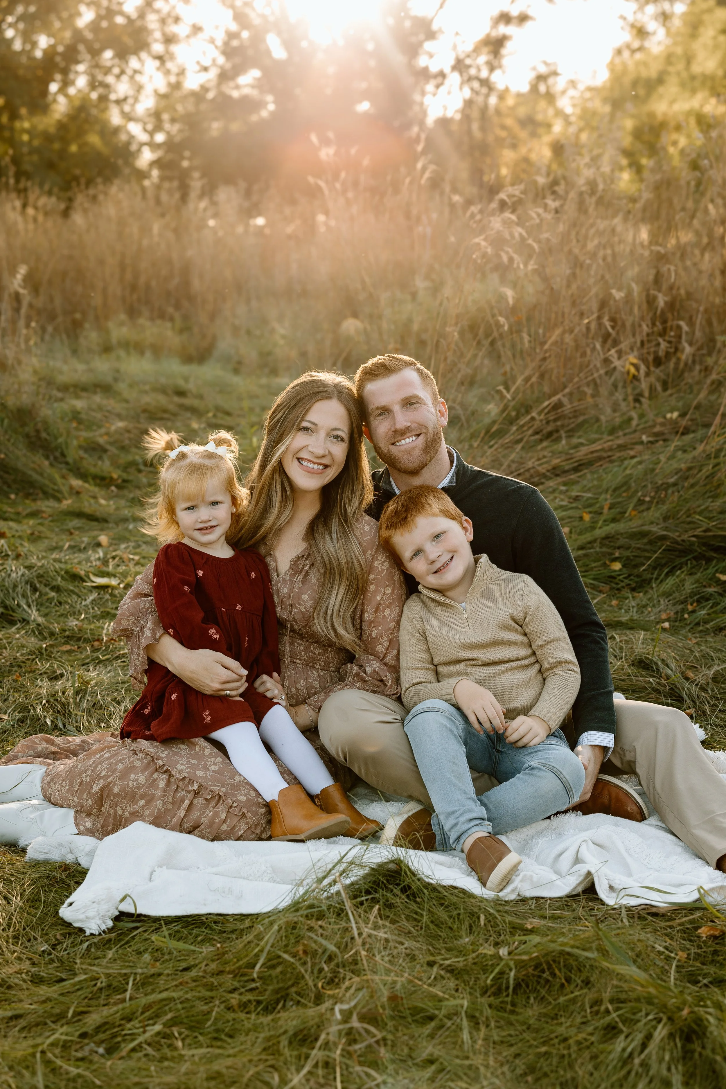Family of four sitting on a blanket in a grassy field during sunset, smiling at the camera. The family includes a mother, father, young girl, and young boy. The background features tall grass and trees.