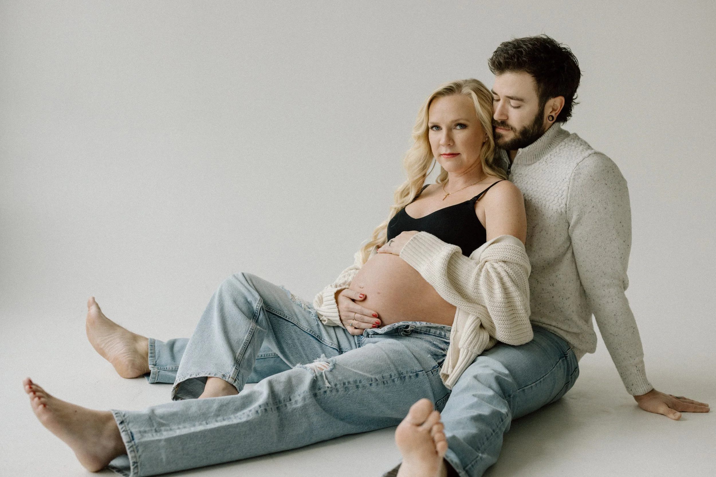 A pregnant woman sitting on a man's lap, with both sitting on the floor against a plain white background. The woman is touching her belly and looking at the camera, while the man is leaning in close with his eyes closed.