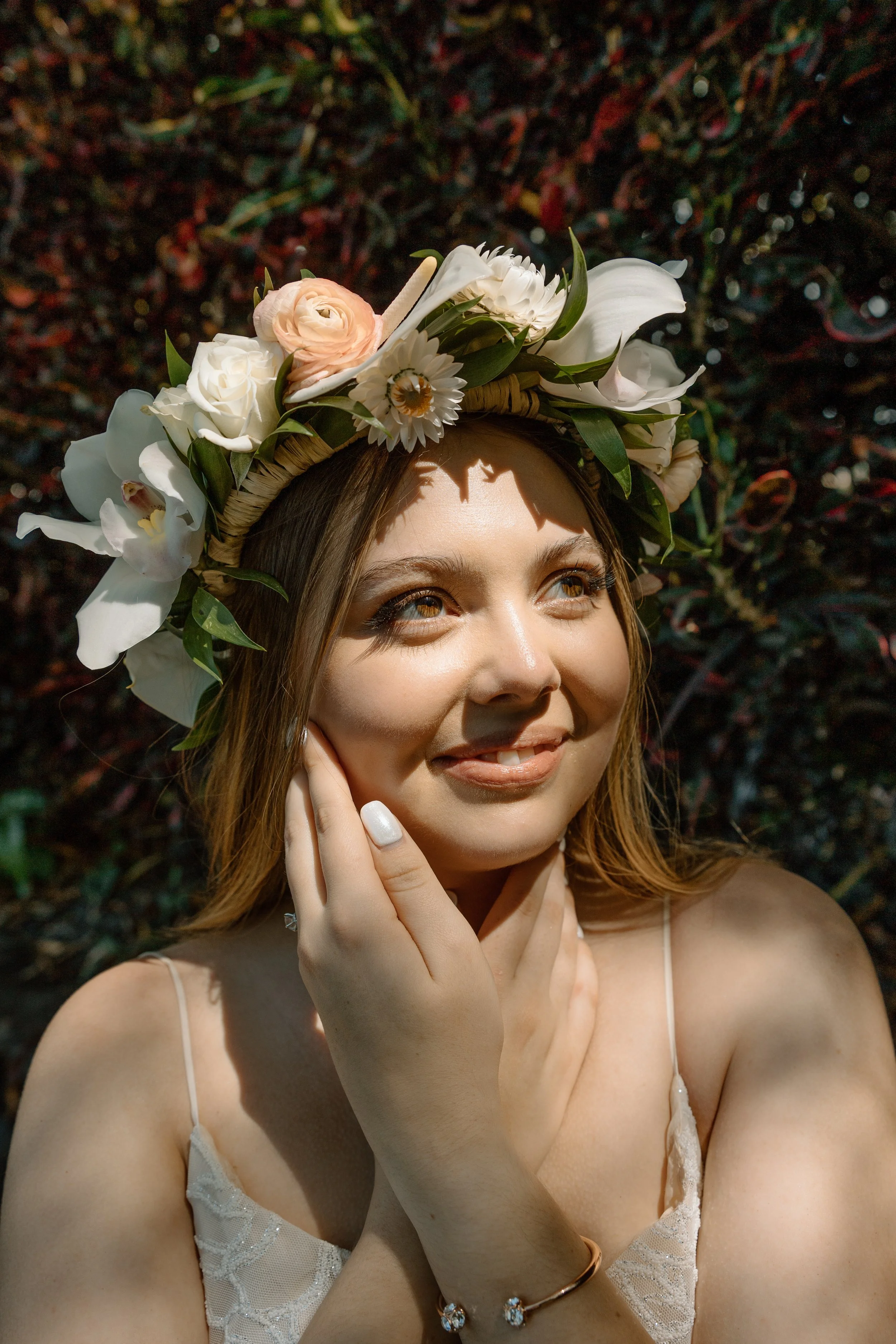 Close-up of a smiling woman wearing a floral crown and a white dress, touching her face, outdoors with sunlight.