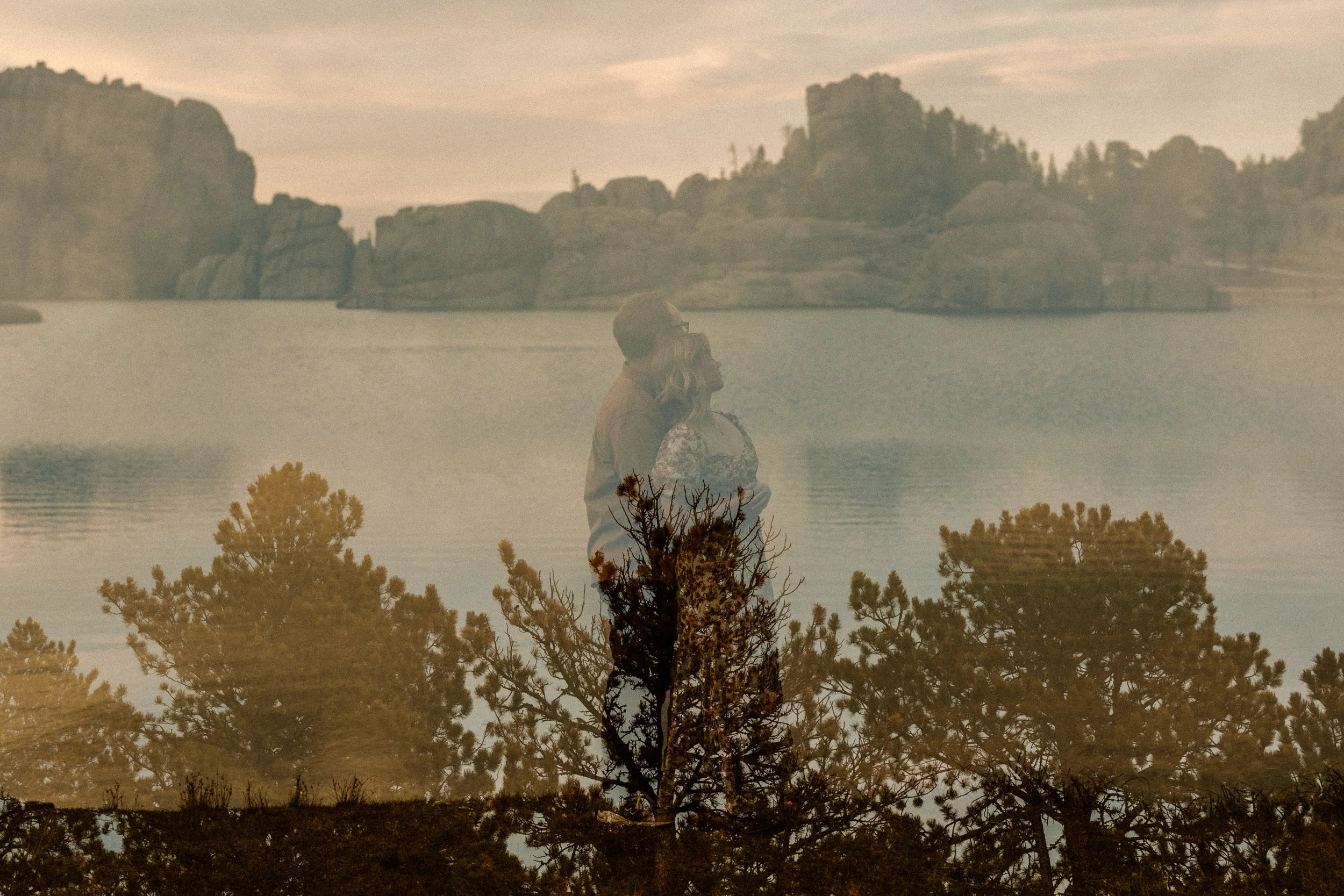 Double exposure photograph of a couple hugging by a lake, with trees and rocky formations in the background.