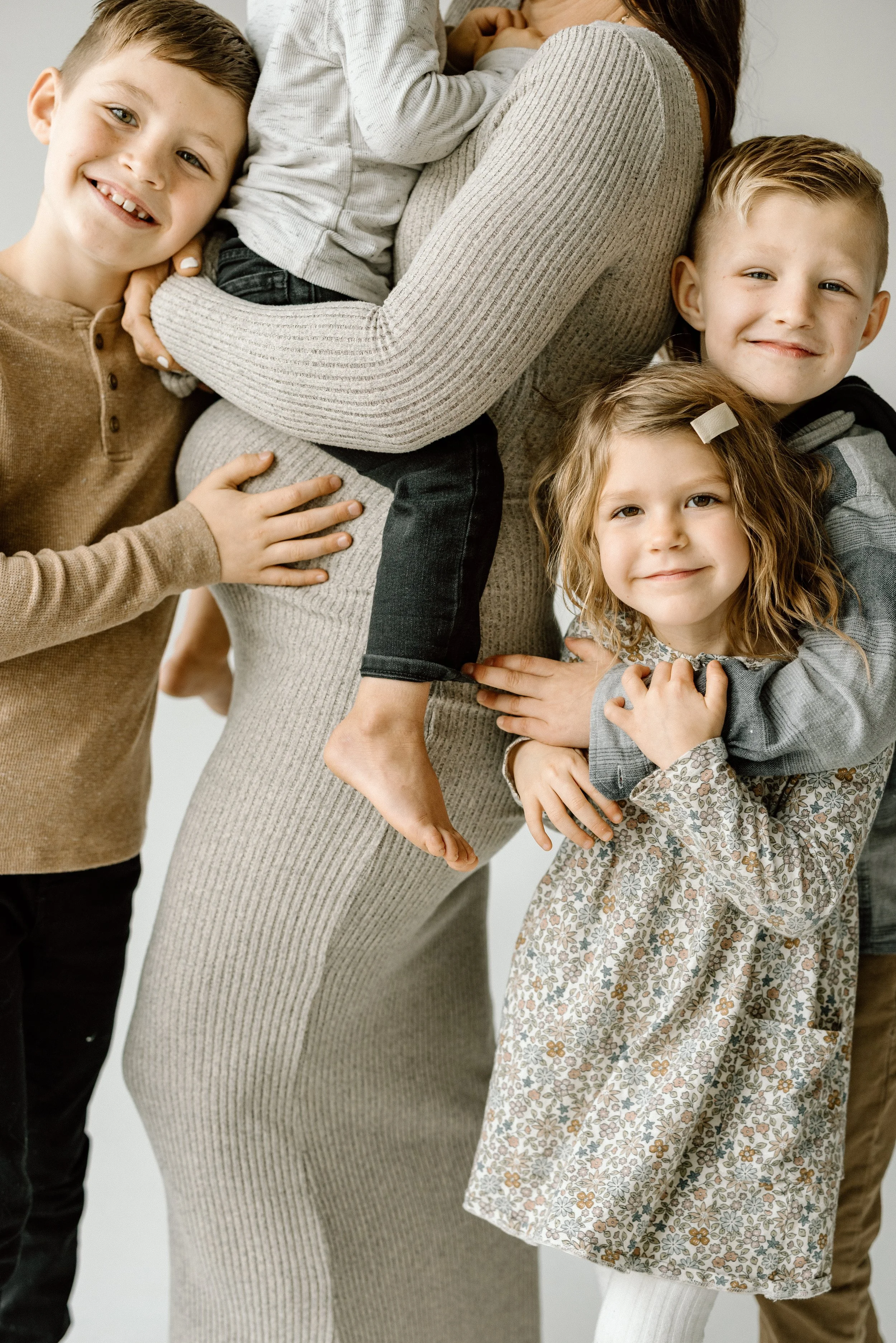 A woman surrounded by four children, hugging and smiling, in a cozy, family portrait setting.