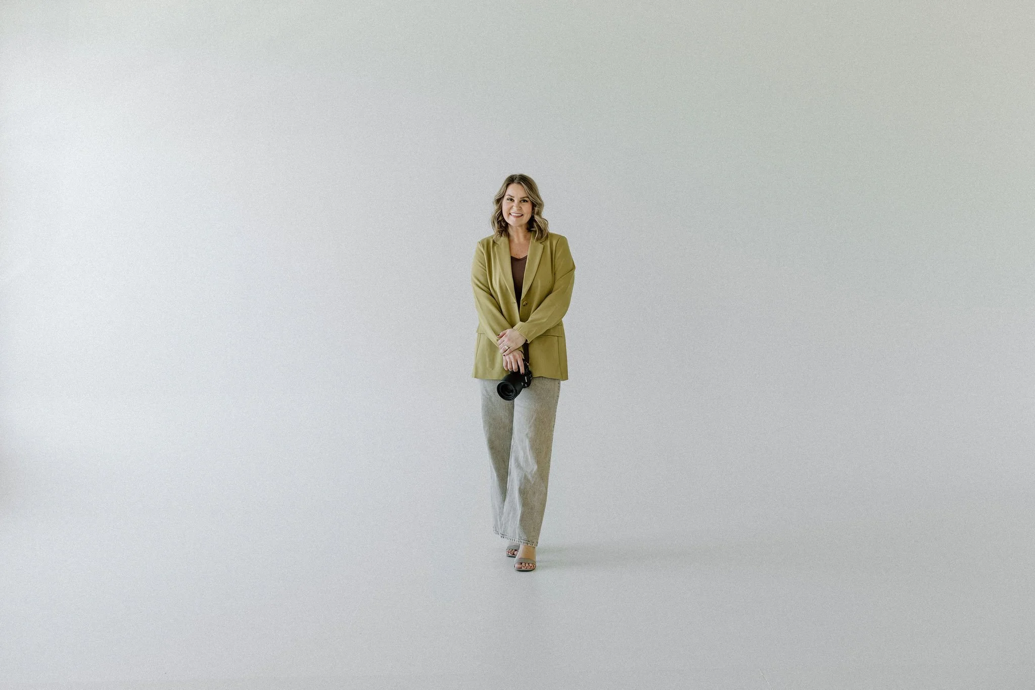 A woman standing in a minimal white studio, holding a camera, smiling, wearing a green blazer and light-colored pants.