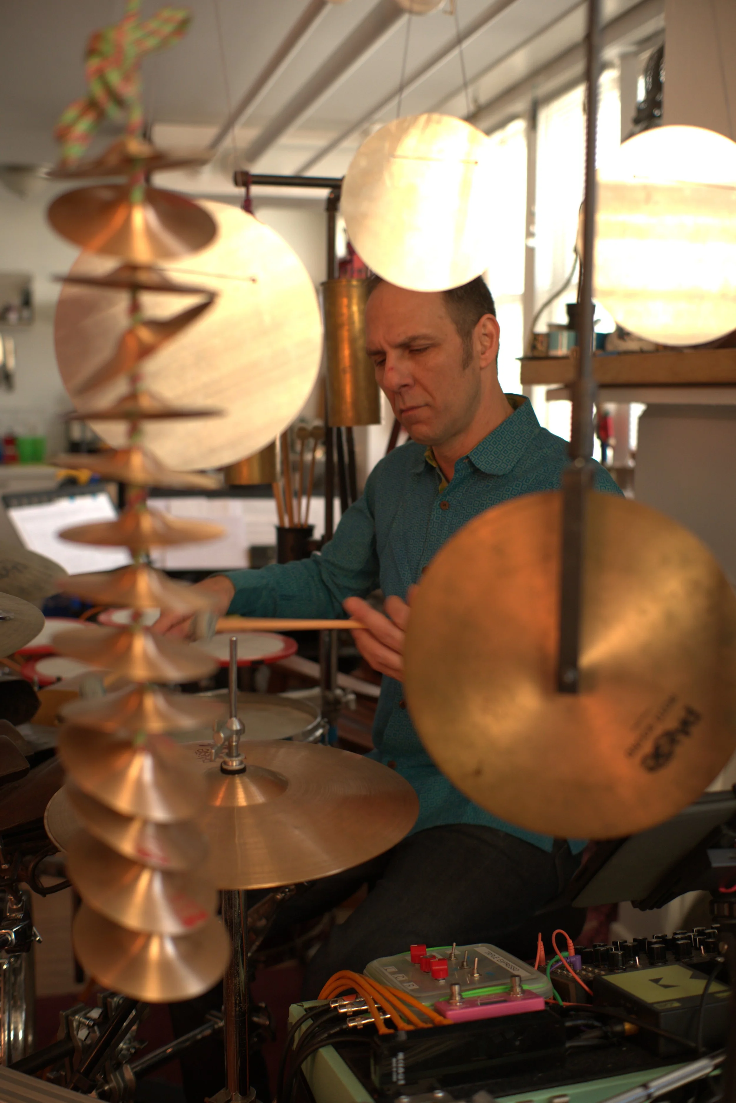 A man is in a music studio, surrounded by musical instruments like cymbals and various electronic devices. He appears focused while working with music equipment.