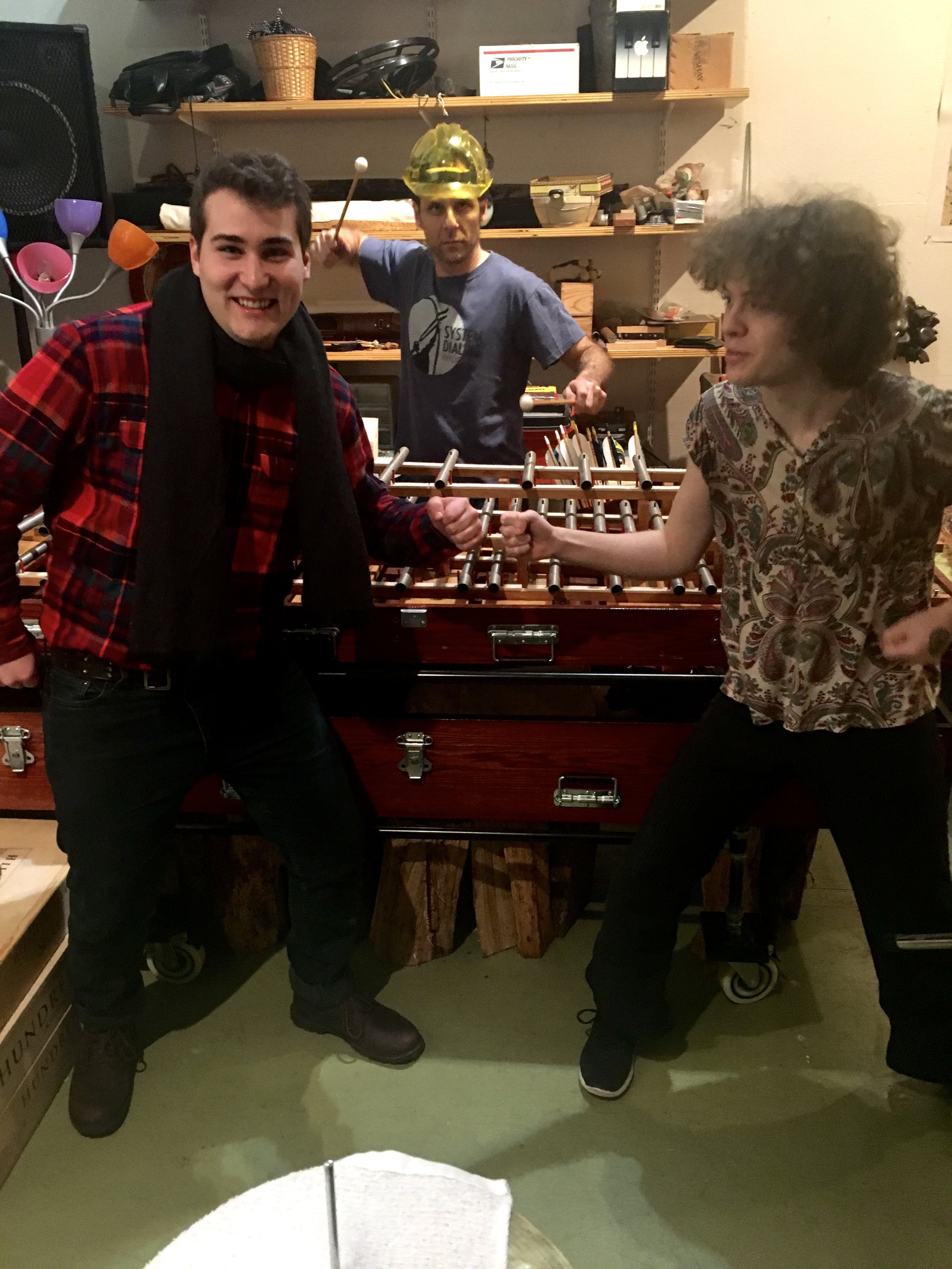 Two young men are standing in front of a foosball table, fist bumping and smiling. One is wearing a red plaid shirt, black scarf, and brown boots; the other is wearing a patterned shirt, black pants, and sneakers. In the background, a third man is wearing a construction helmet and a gray T-shirt, holding a mallet, with shelves filled with various items behind him.