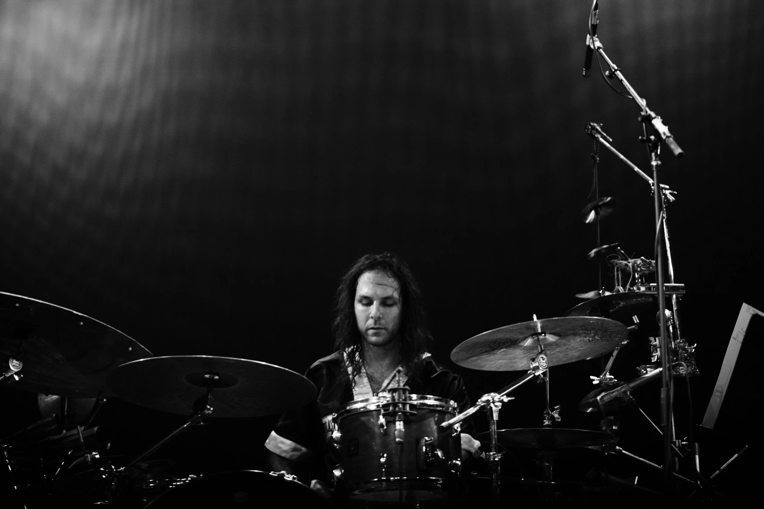 A black and white photo of a man with long hair playing drums on stage, with microphones and drum equipment visible.