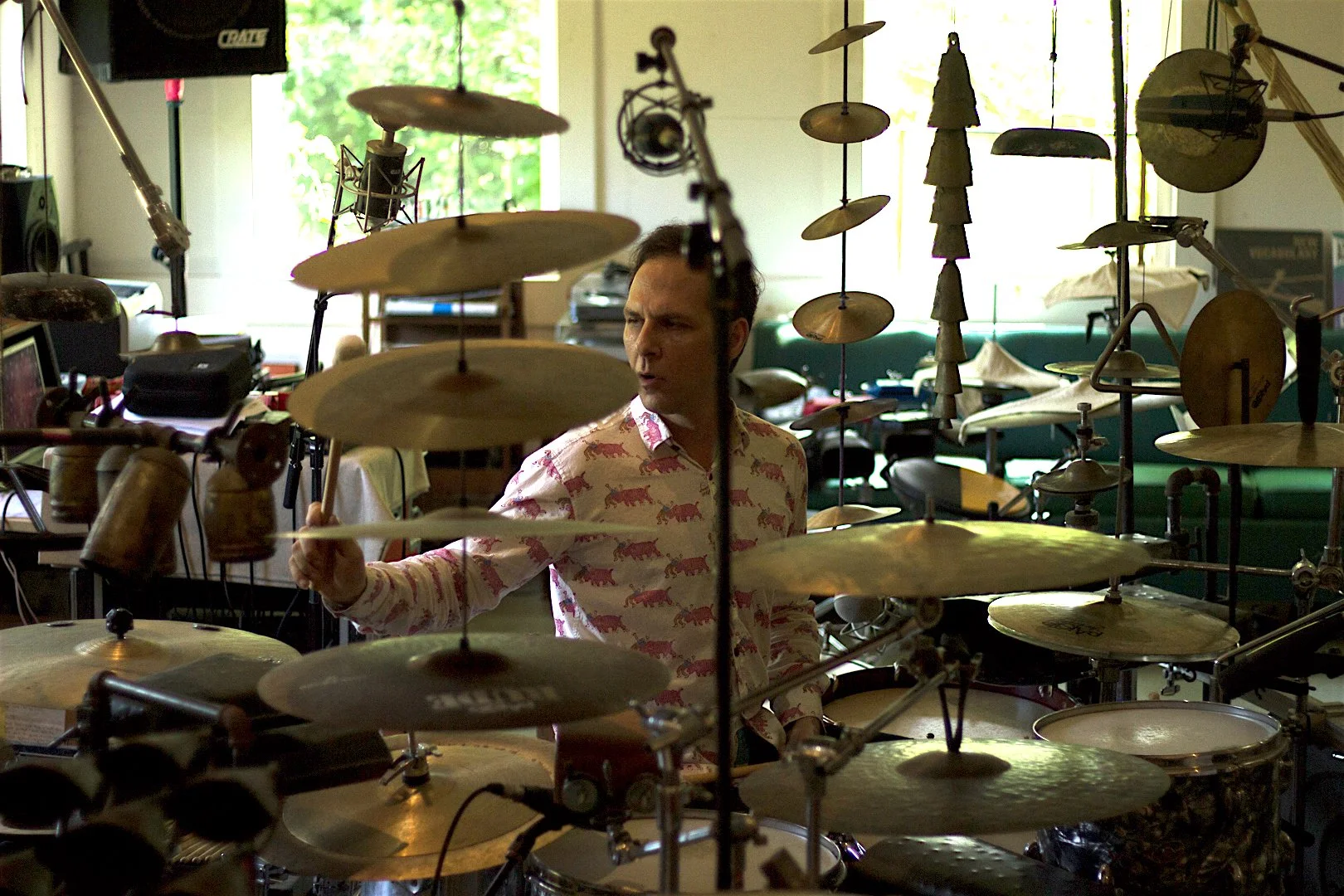 A person sitting behind an extensive drum set, wearing a shirt with a pink panther pattern, inside a room with musical equipment and large windows with natural light.