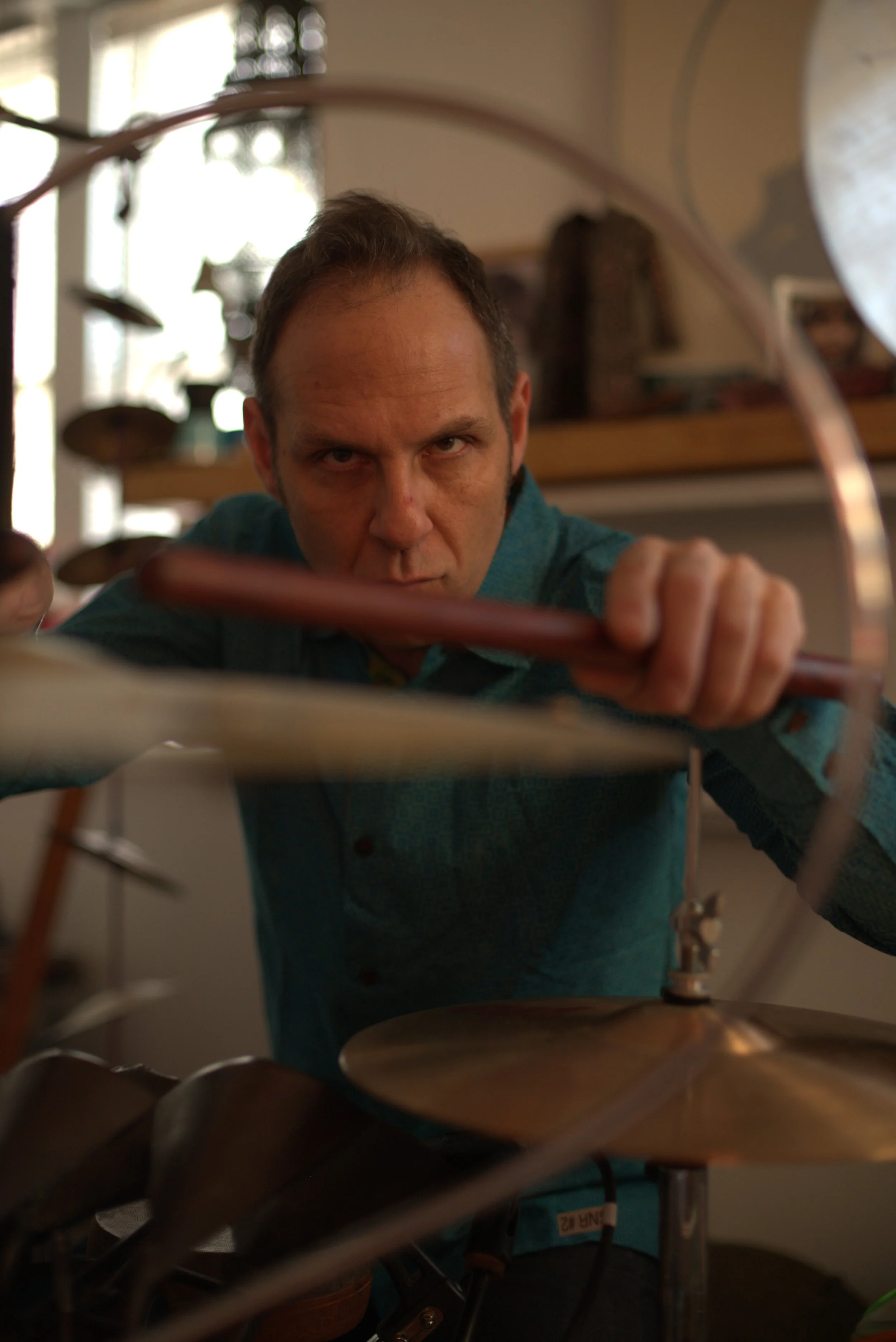 A man with a focused expression reaching towards the camera, seen through a metallic rack with round objects, possibly cymbals, in a room with shelves and windows in the background.