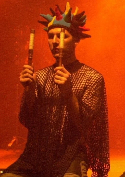 Person wearing a multicolored, spiked costume headpiece and playing two recorders, dressed in a fishnet shirt against an orange background.