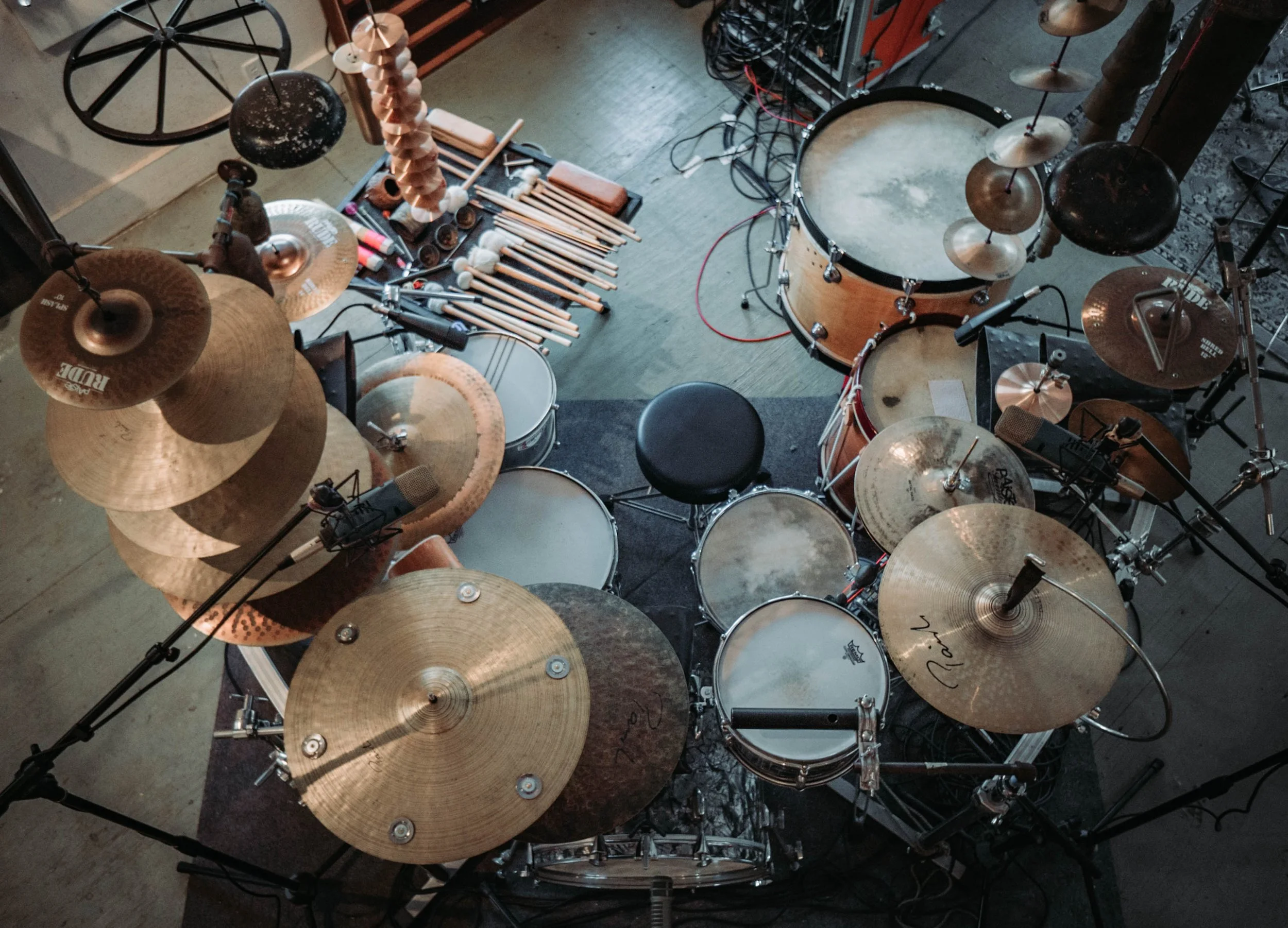 Overhead view of a drum set with multiple cymbals, drumsticks, and percussion instruments arranged on a wooden floor.