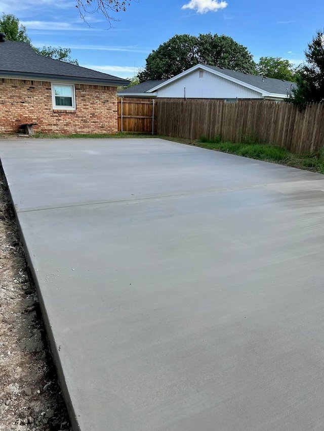 Freshly poured concrete driveway next to a brick house and a wooden fence, with a house and trees in the background.