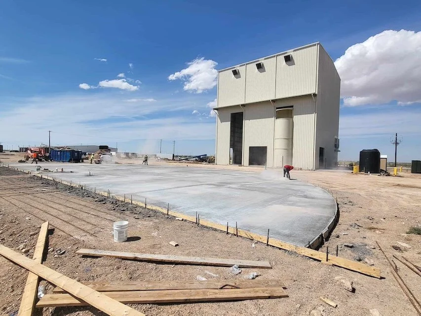 Construction site with a large industrial building, workers smoothing concrete on the ground, and construction materials scattered around. Clear blue sky with some clouds.