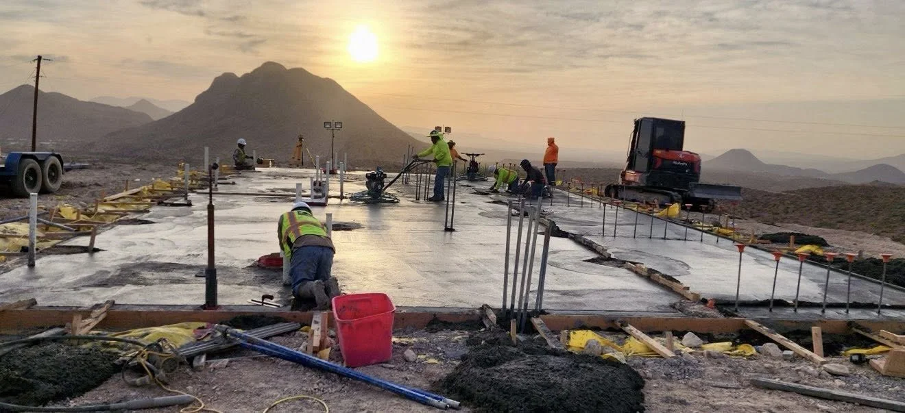 Construction workers laying concrete on a flat surface during sunset, with mountains in the background and construction equipment around.