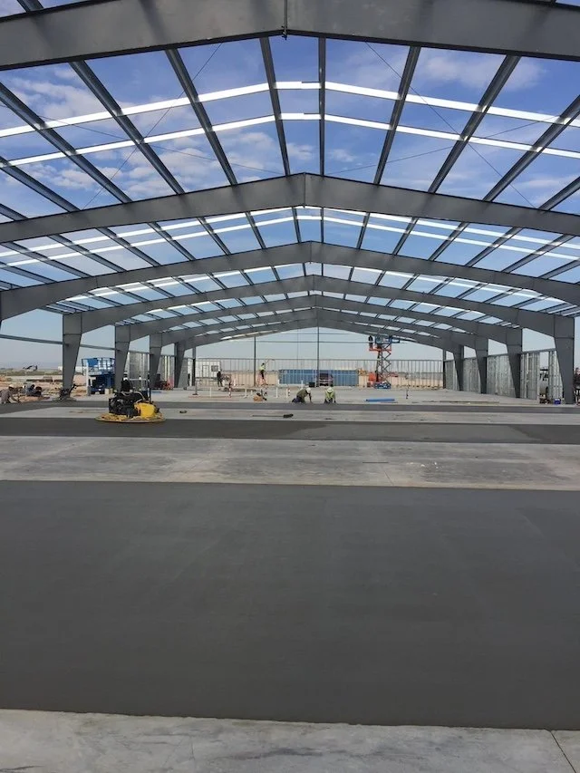 Construction of a large glass and metal canopy structure at an airport or similar facility, with workers and construction equipment visible.