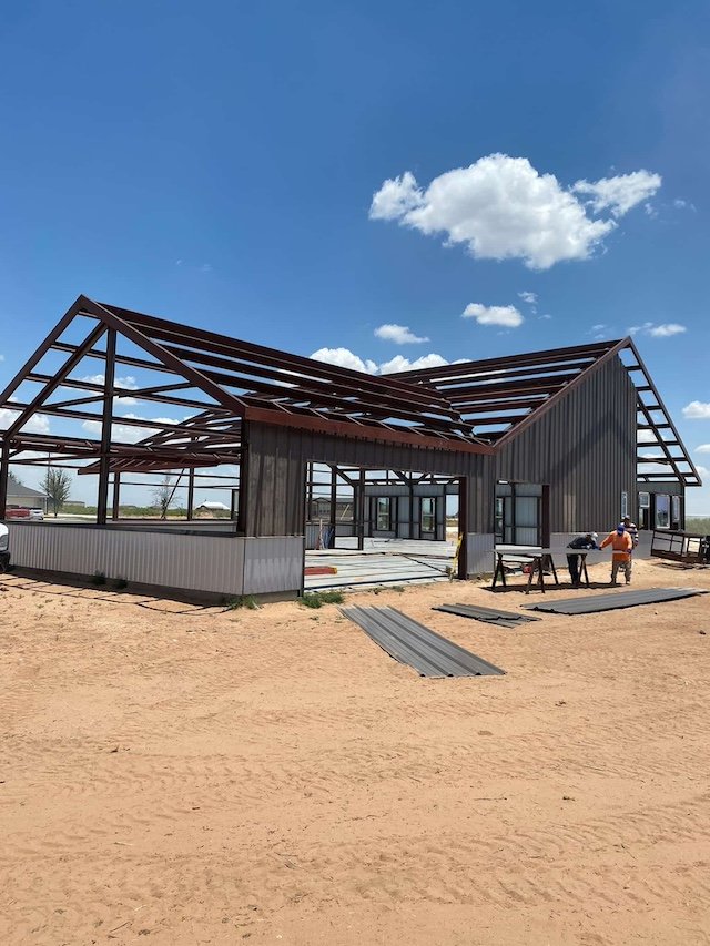 Under construction house with steel roof frame and brown siding, set on a sandy lot with workers and tools, under a blue sky with clouds.