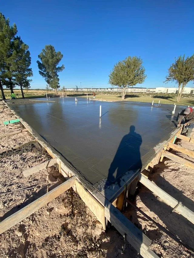 Freshly poured concrete foundation for a building, surrounded by wooden formwork and rebar, with trees and a clear blue sky in the background.