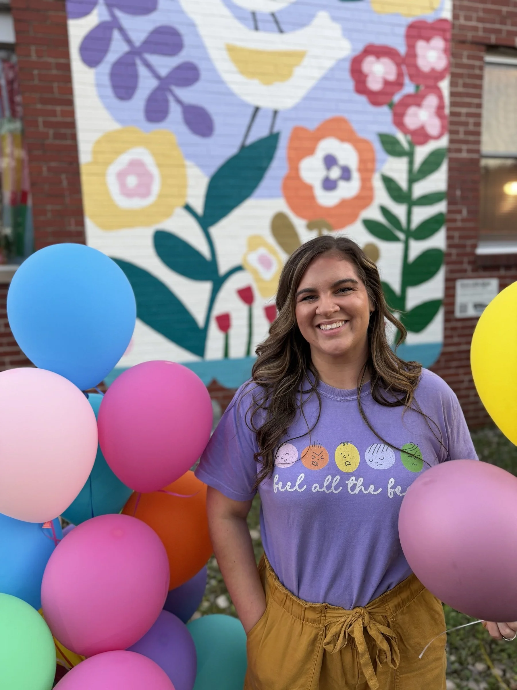 Smiling Sandbox therapist holding colorful balloons and wearing the violet "feel all the feels" shirt