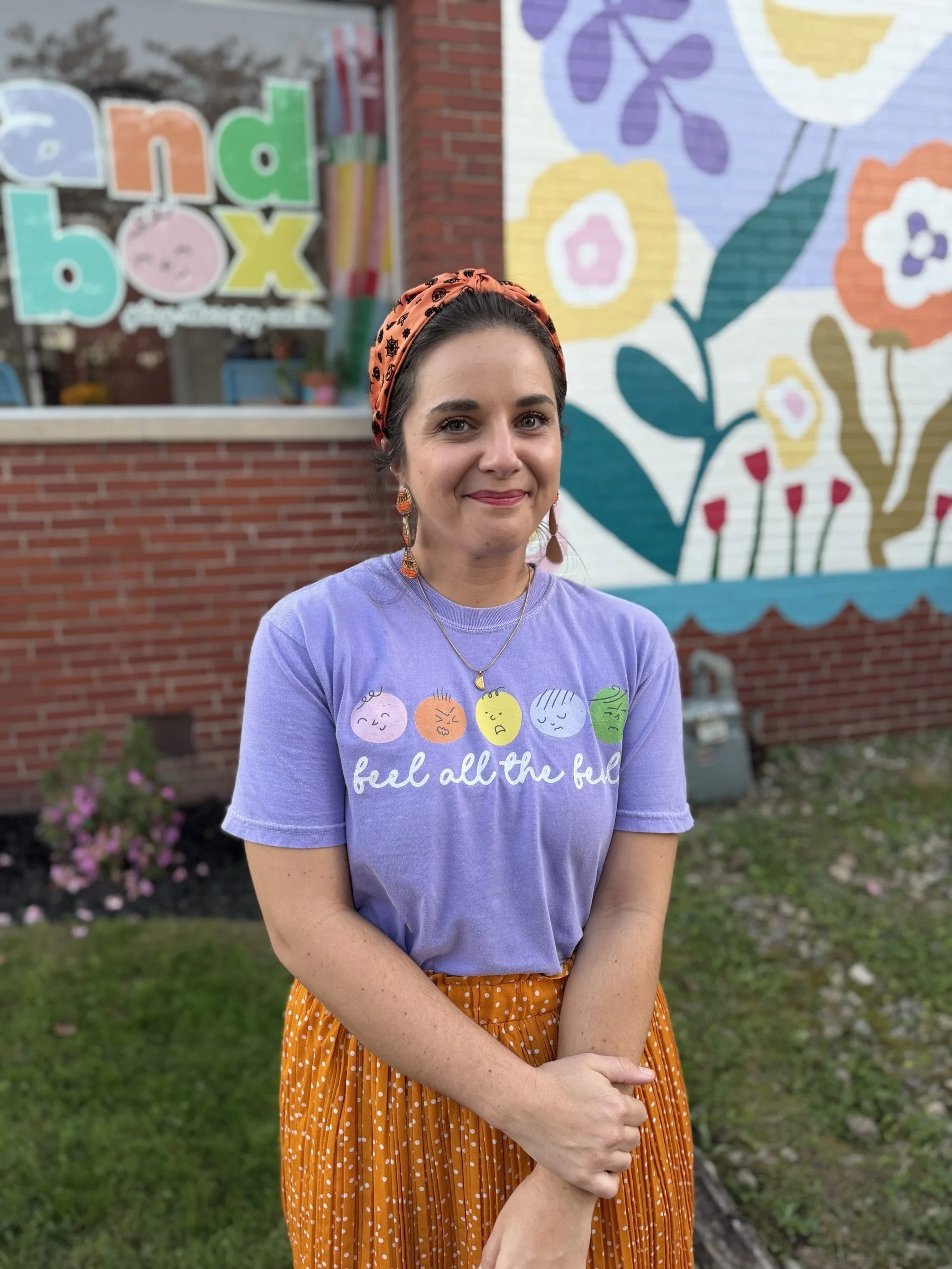 Sandbox therapist in an orange headband wearing the violet "feel all the feels" shirt in front of the Sandbox flower mural
