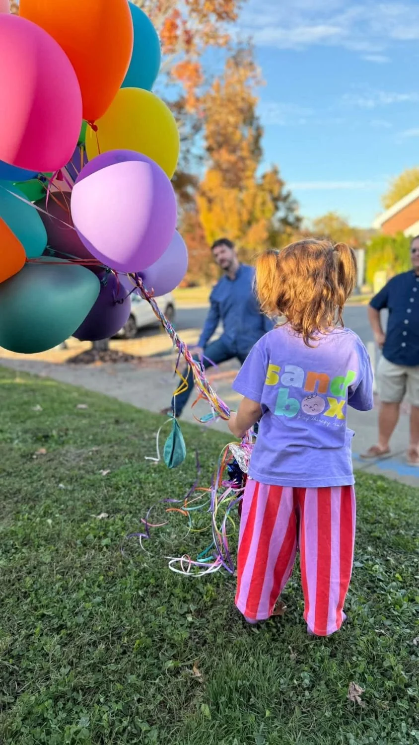 Back of the violet Sandbox shirt with the colorful Sandbox Play Therapy Center logo, worn by a child holding balloons