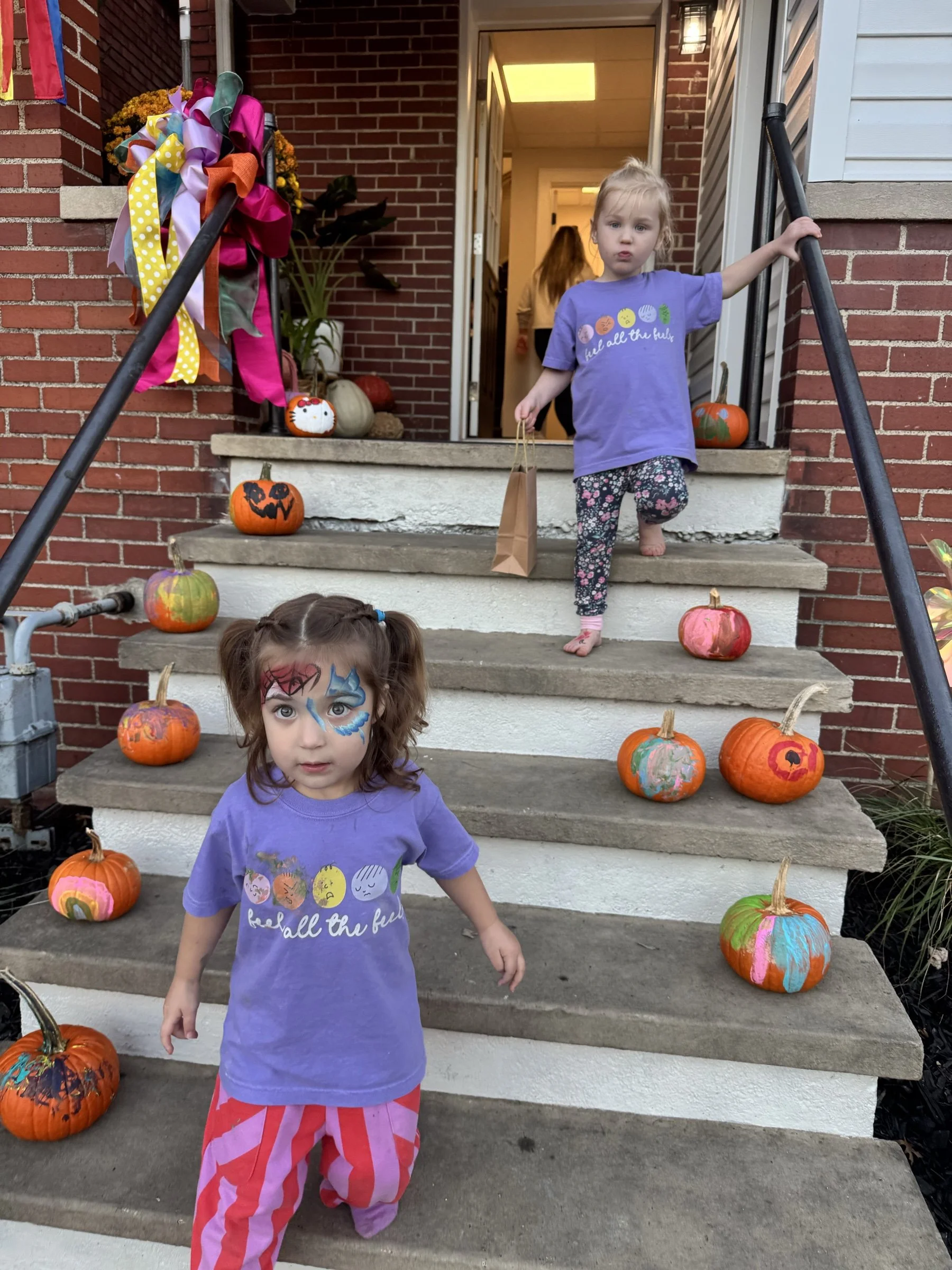 Two young girls wearing youth "feel all the feels" shirts on porch steps lined with painted pumpkins