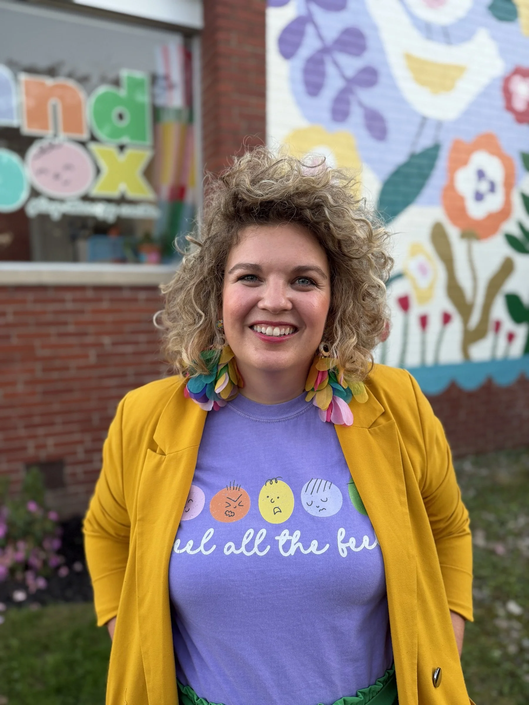 Smiling Sandbox therapist with curly hair in a yellow blazer wearing the violet "feel all the feels" shirt