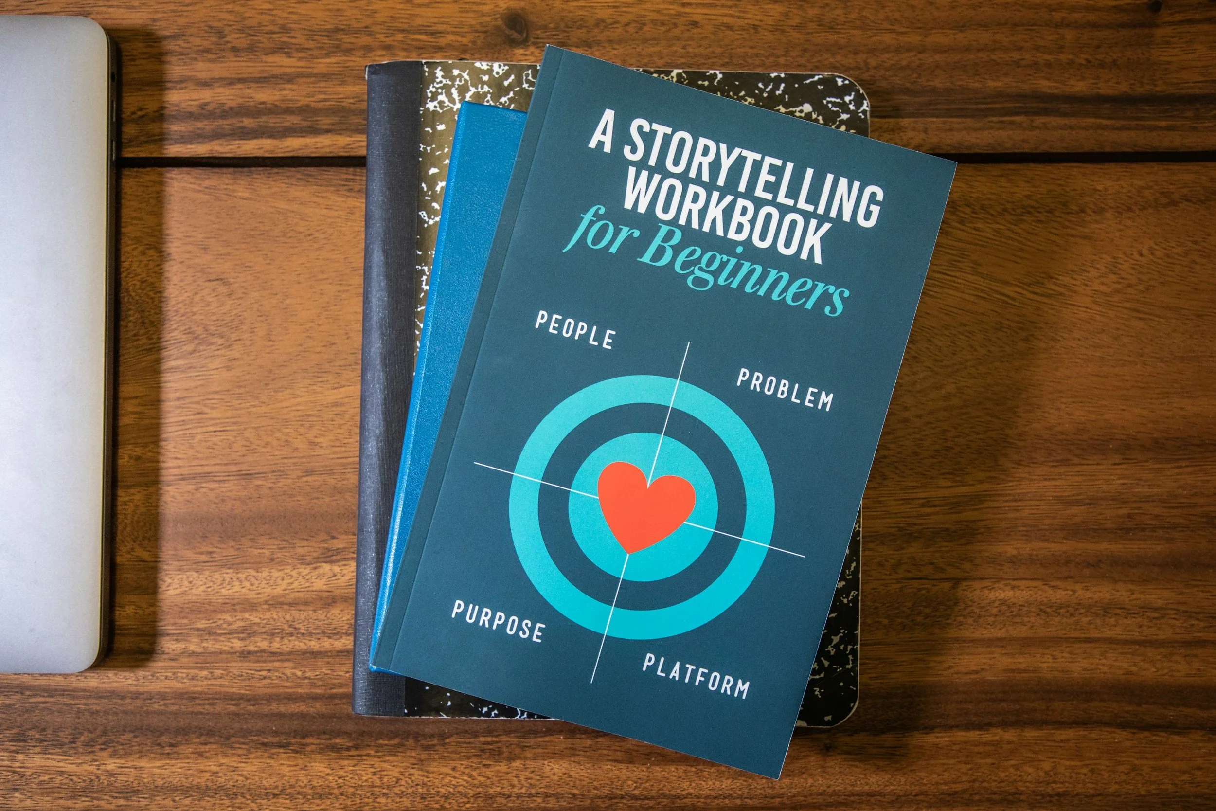 A blue book titled 'A Storytelling Workbook for Beginners' resting on top of a black and white notebook, which is placed on top of a closed laptop on a wooden surface.