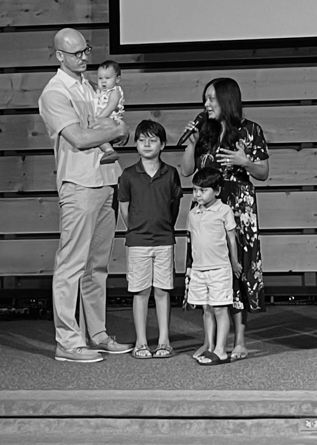 A man holding a young girl on his arm next to a woman speaking into a microphone, with two boys standing in front of them, inside a room with wooden paneling.