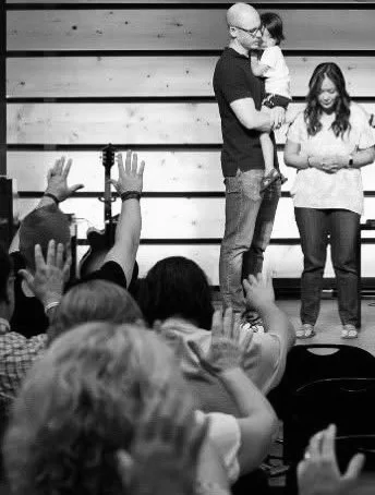 A man holding a girl on a stage with a woman standing beside them, while several children in the audience raise their hands.