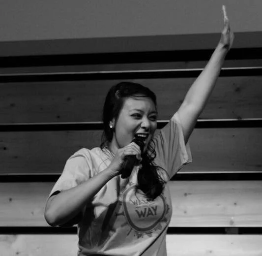 A woman speaking into a microphone with one arm raised, smiling, in a casual shirt, on a stage with wooden paneling.