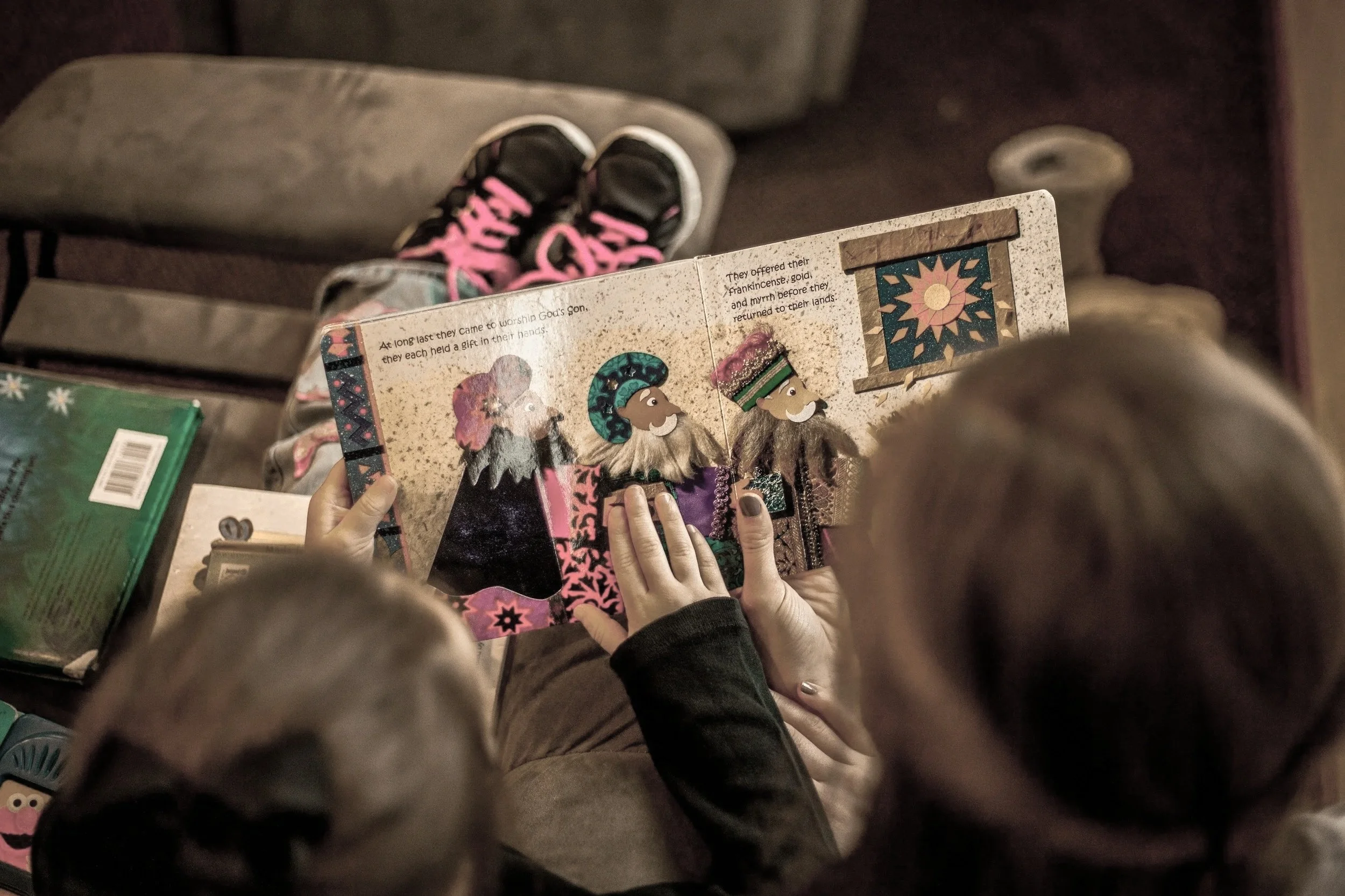 A child and an adult sitting on a couch reading a colorful Christmas storybook featuring illustrations of three bearded wise men wearing colorful robes and crowns.