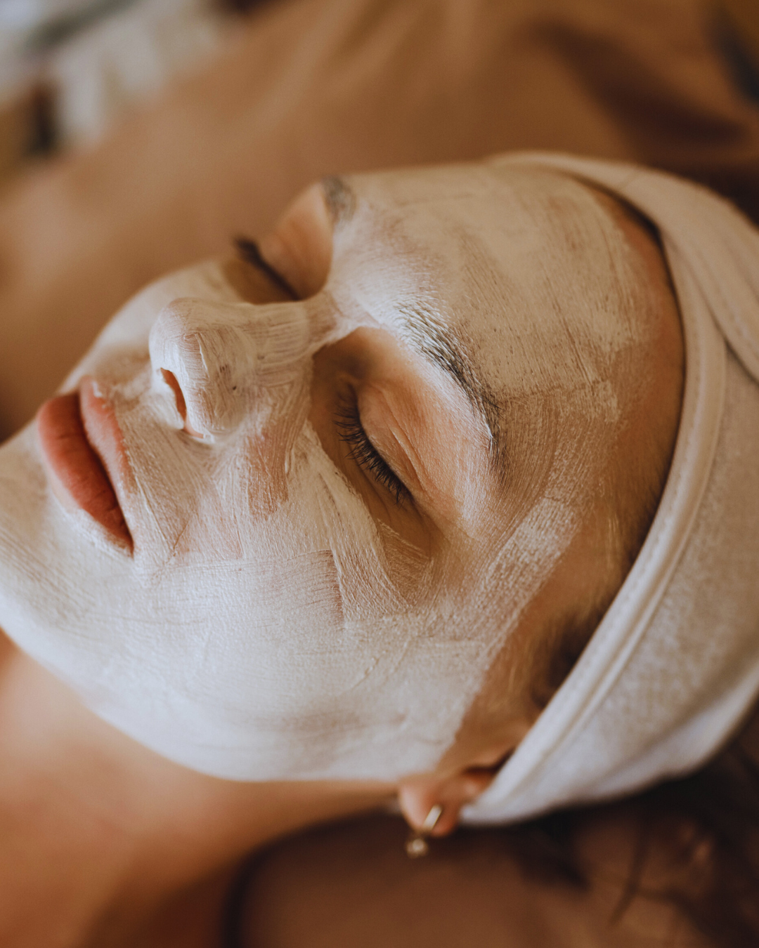 A woman lying down with a white facial mask applied to her face, eyes closed, and wearing a headband.