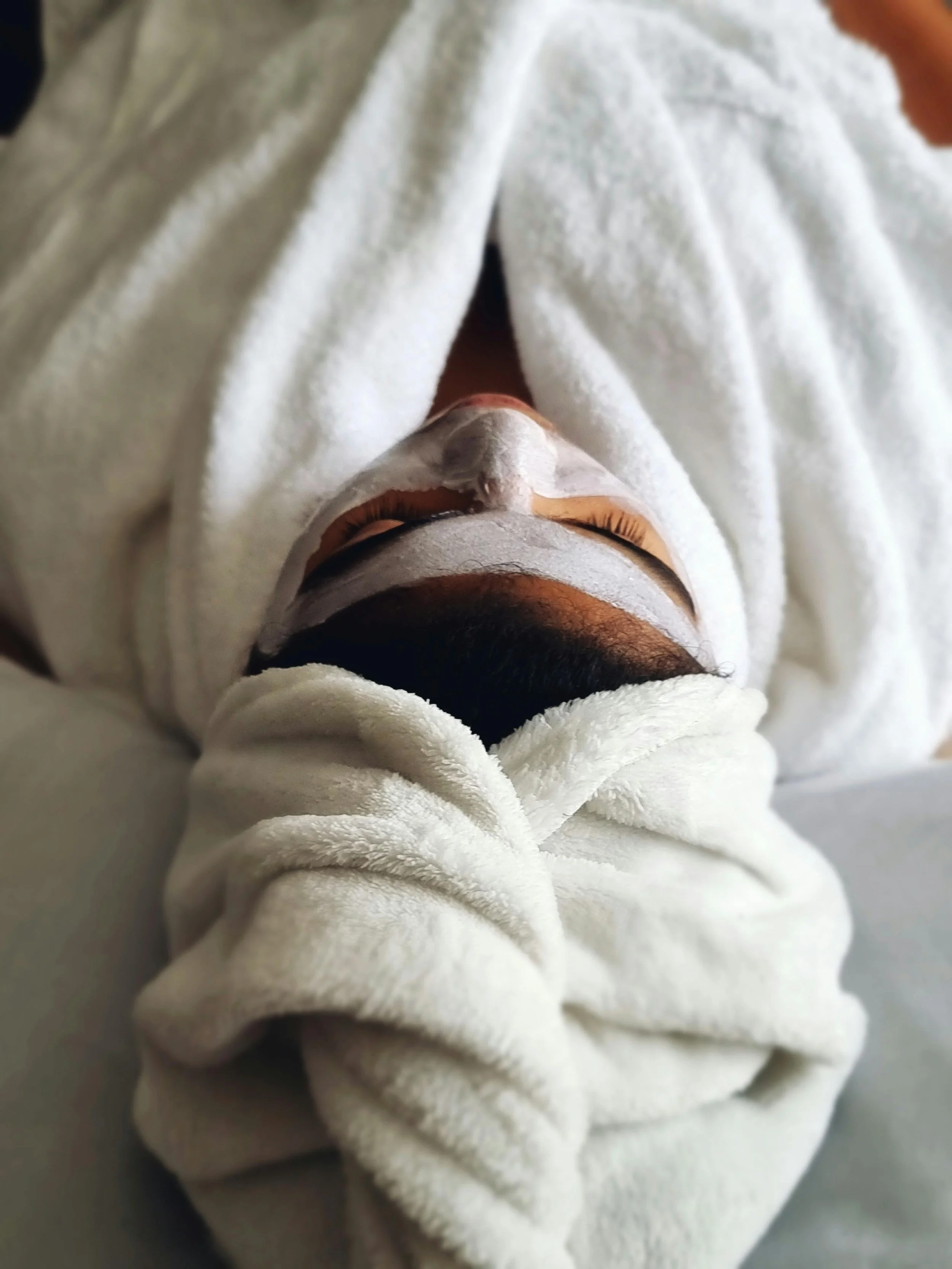 Person lying on a bed with a white towel wrapped around their head, wearing a face mask, and dressed in a white robe.