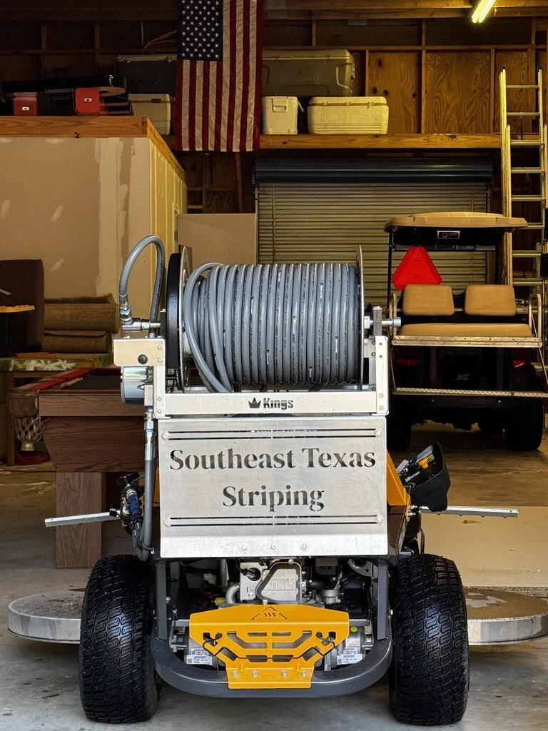 An industrial pressure washer with a large hose reel in a garage, with an American flag hanging above, and various storage boxes and a golf cart in the background.