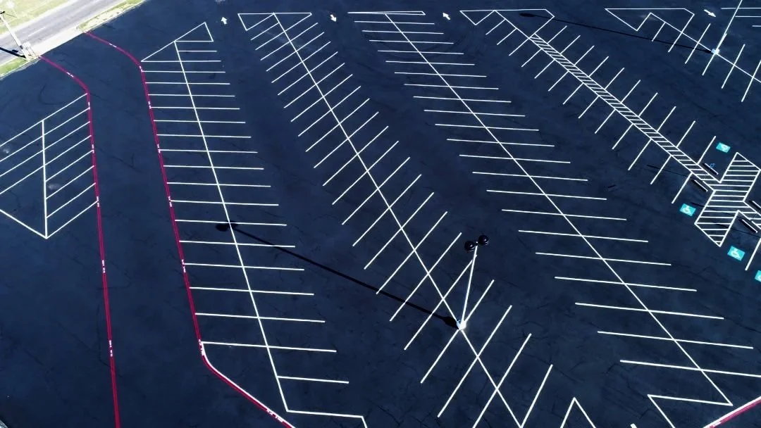 An empty parking lot with white parking space lines, a few blue marked handicap spots, and a small landscaped area at the top of the image.