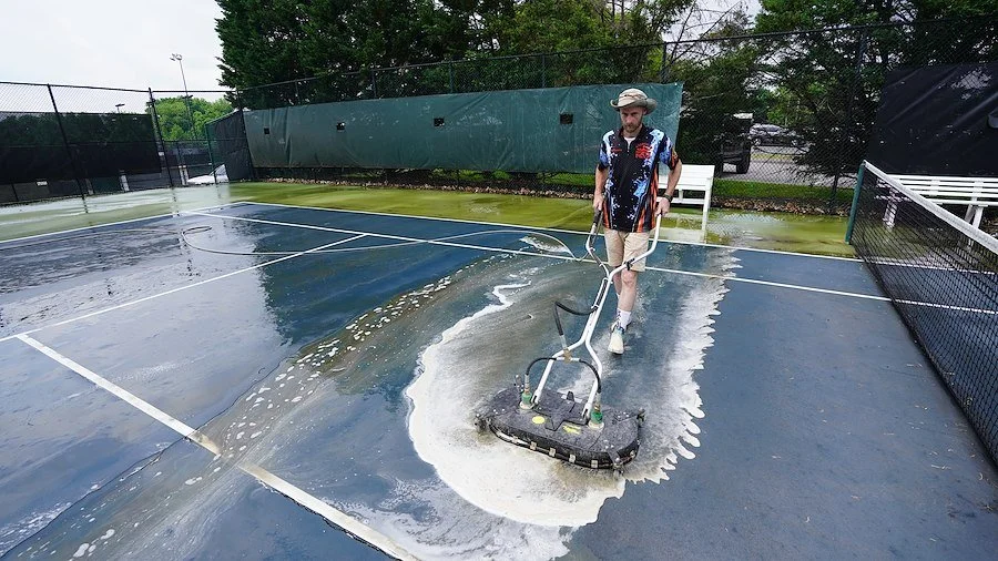 Person cleaning a wet tennis court with a floor scrubber, causing water to splash.