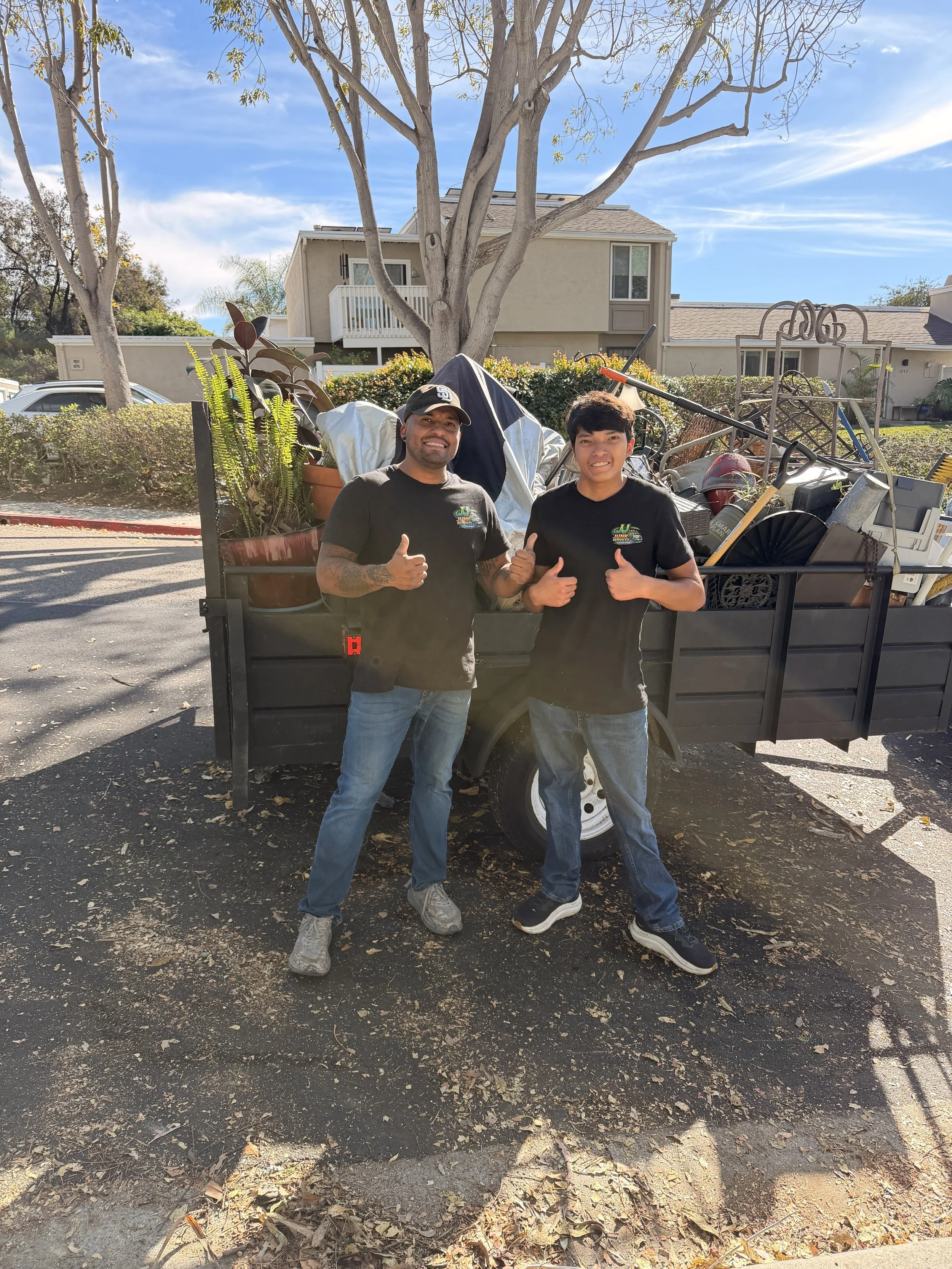 Two men standing in front of a trailer filled with yard debris and plants, giving thumbs-up signs, outdoors on a sunny day with houses and trees in the background.