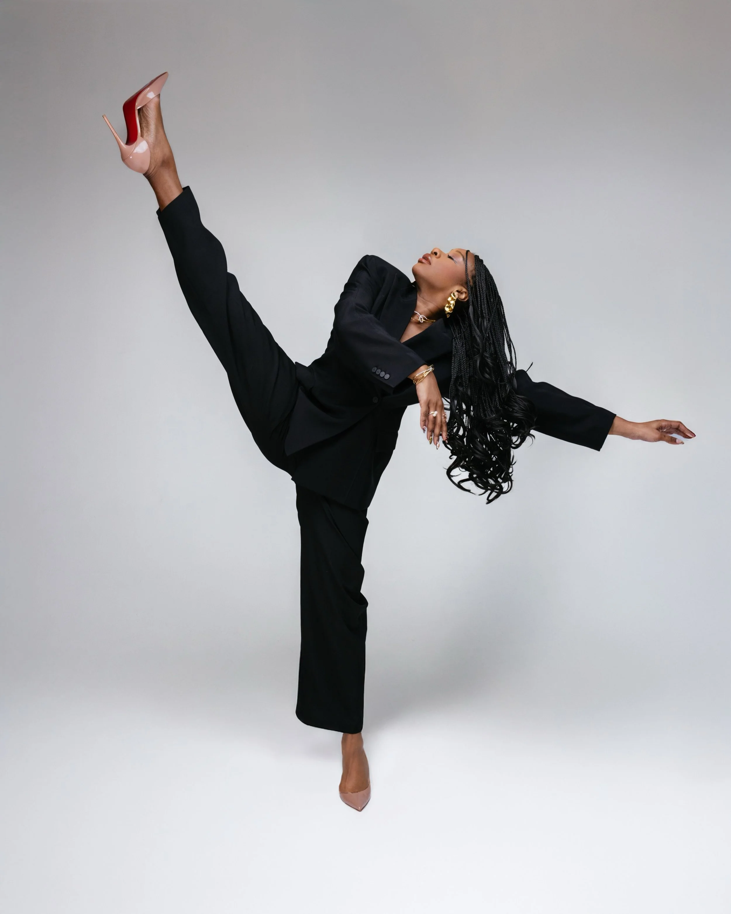 Woman in black suit performing a high kick, wearing nude heels and gold jewelry, with long braids and a neutral background.