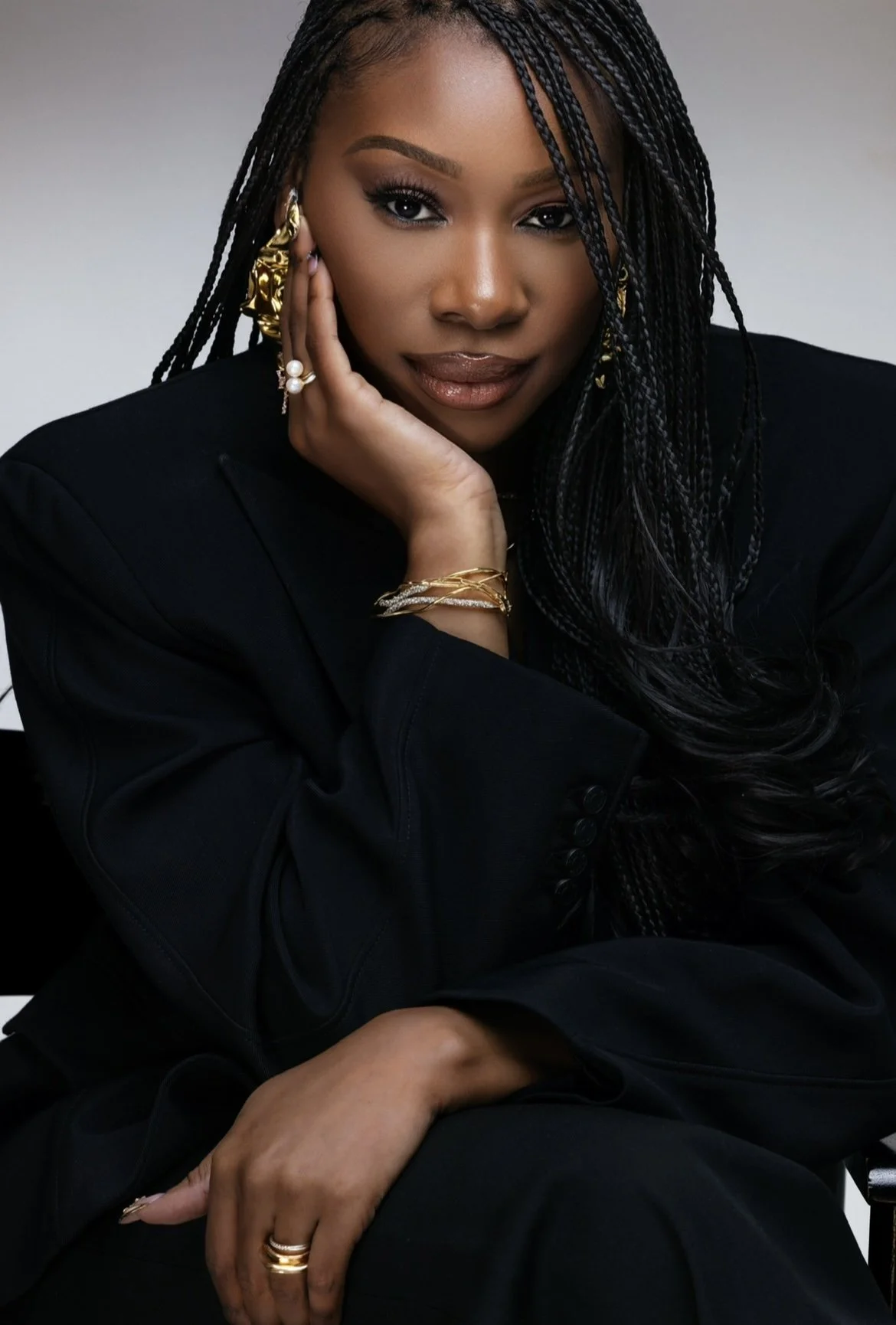 A woman with dark braided hair, wearing a black blazer, gold jewelry, and earrings, poses with her hand resting on her face against a plain neutral background.
