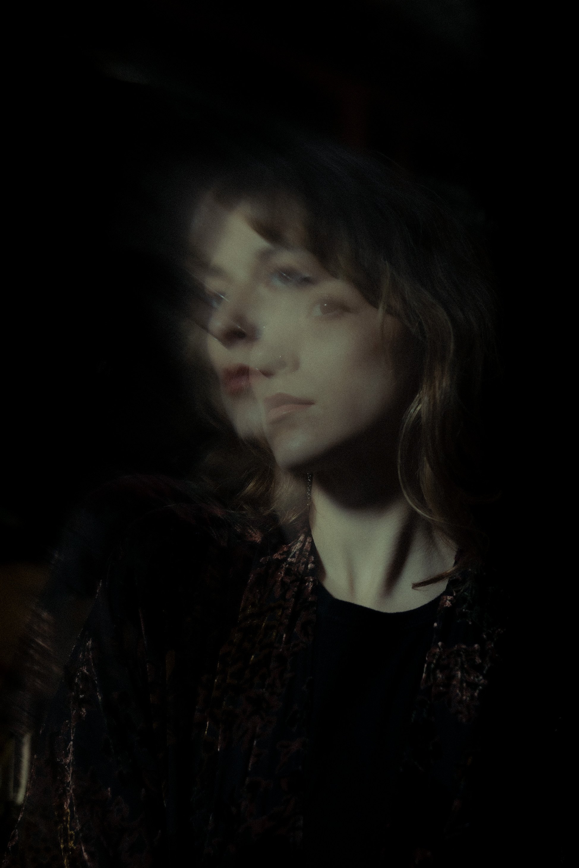 A woman (Emma) (Lead Photographer) with wavy brown hair wearing a velvet kimono, with a motion blur effect making her face appear multiple times against a dark background.