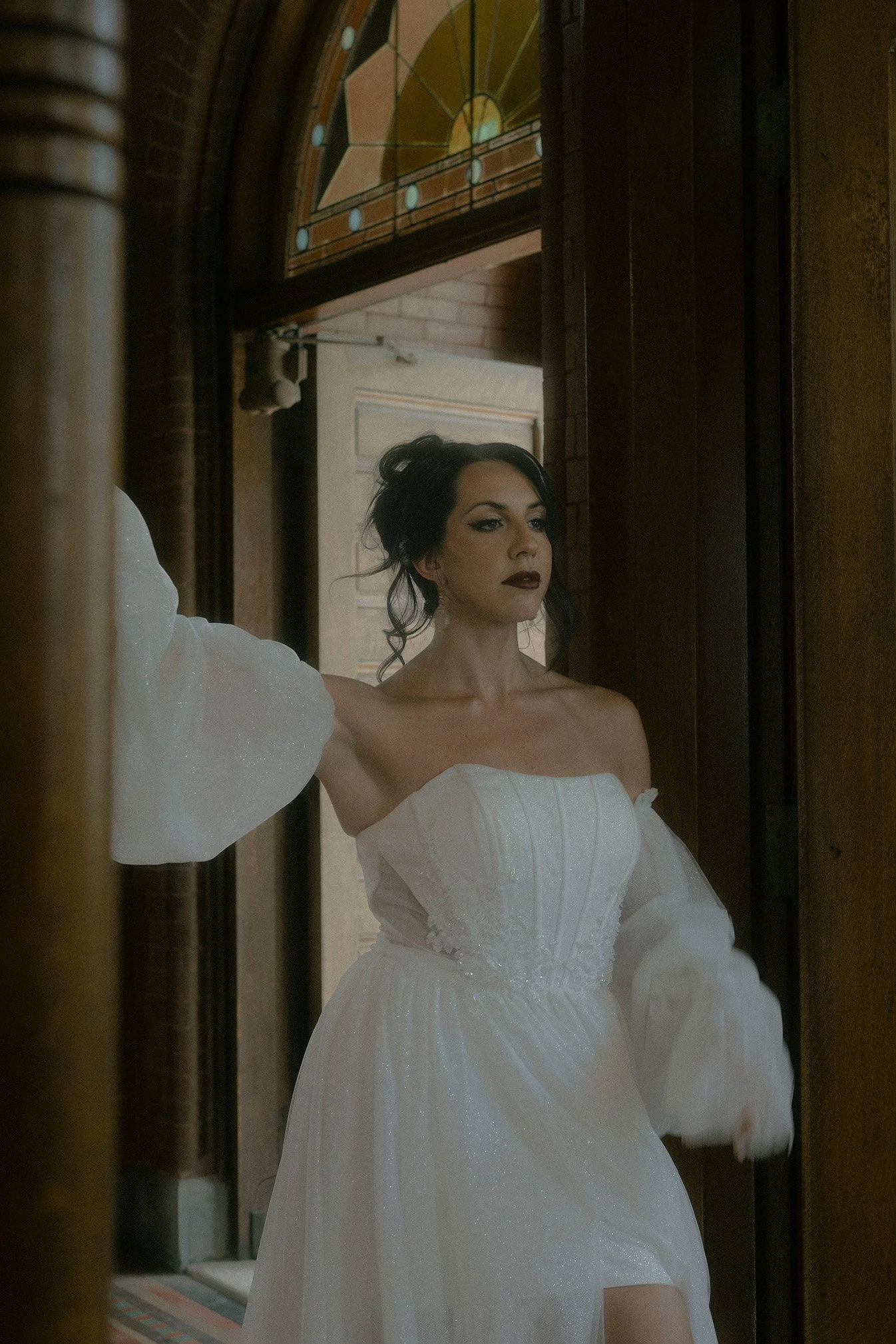 A woman with dark hair and makeup in a white off-shoulder dress standing near a wooden door with stained glass window, looking thoughtfully and confident off camera. 