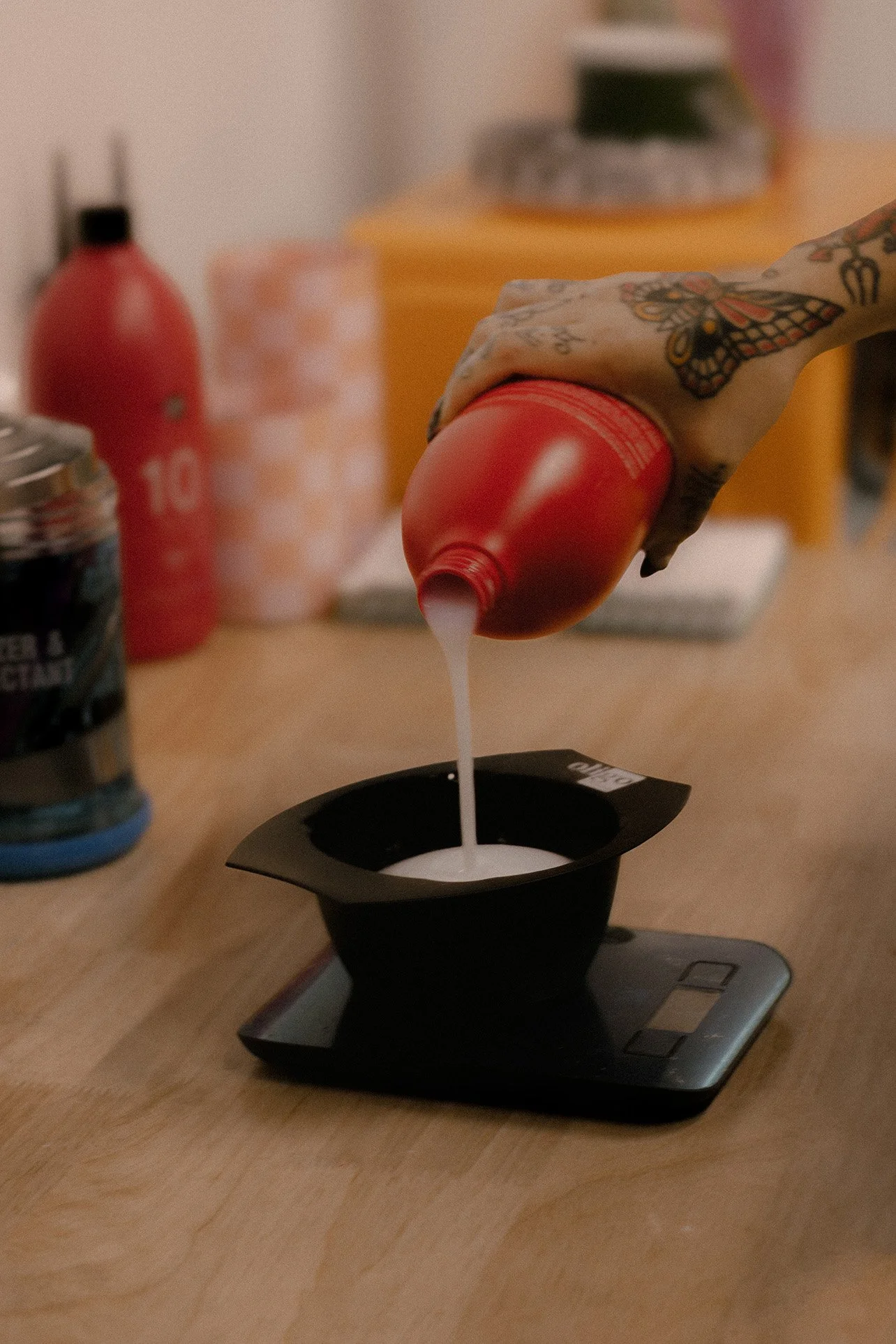 Hair stylist pouring bleach from a red bottle into a black container on a digital kitchen scale on a wooden countertop.