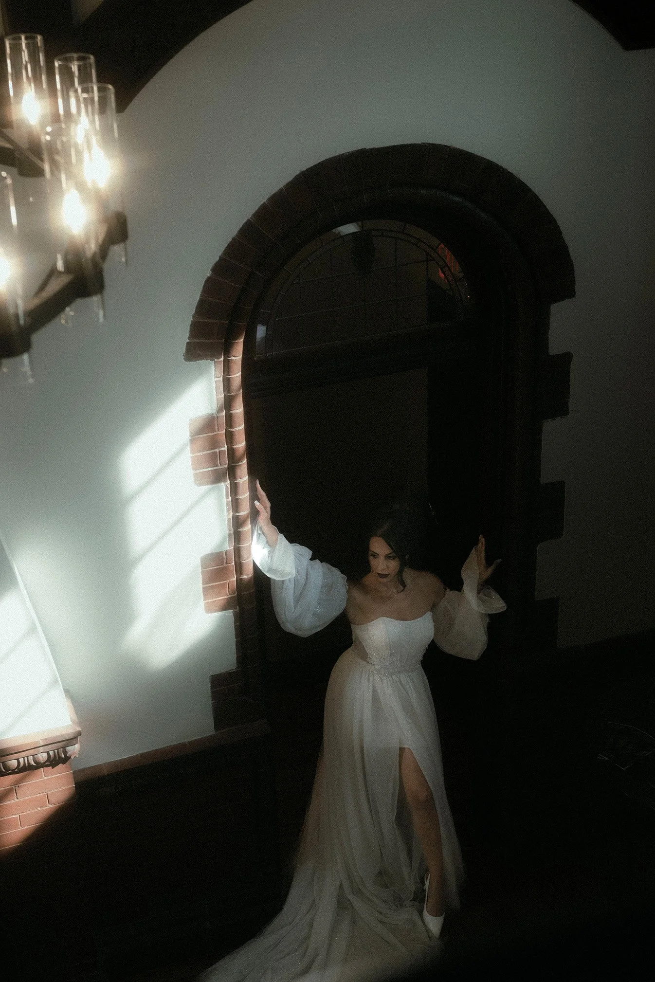 A woman in a white dress with puffy sleeves and a slit in the skirt stands in a dimly lit room, leaning against a brick wall under a window with shadows cast by sunlight.