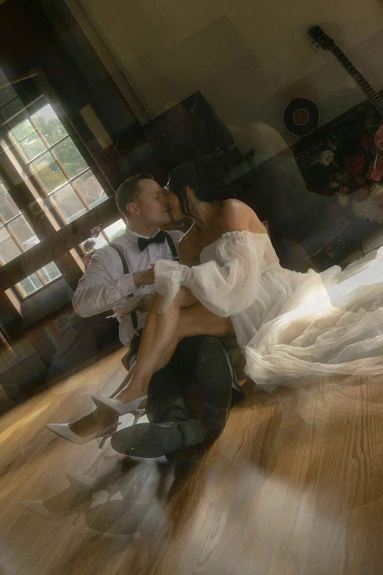 A couple in formal wear, the man in a tuxedo and the woman in a white dress, sharing an intimate moment and kissing inside a room with windows and musical instruments in the background.