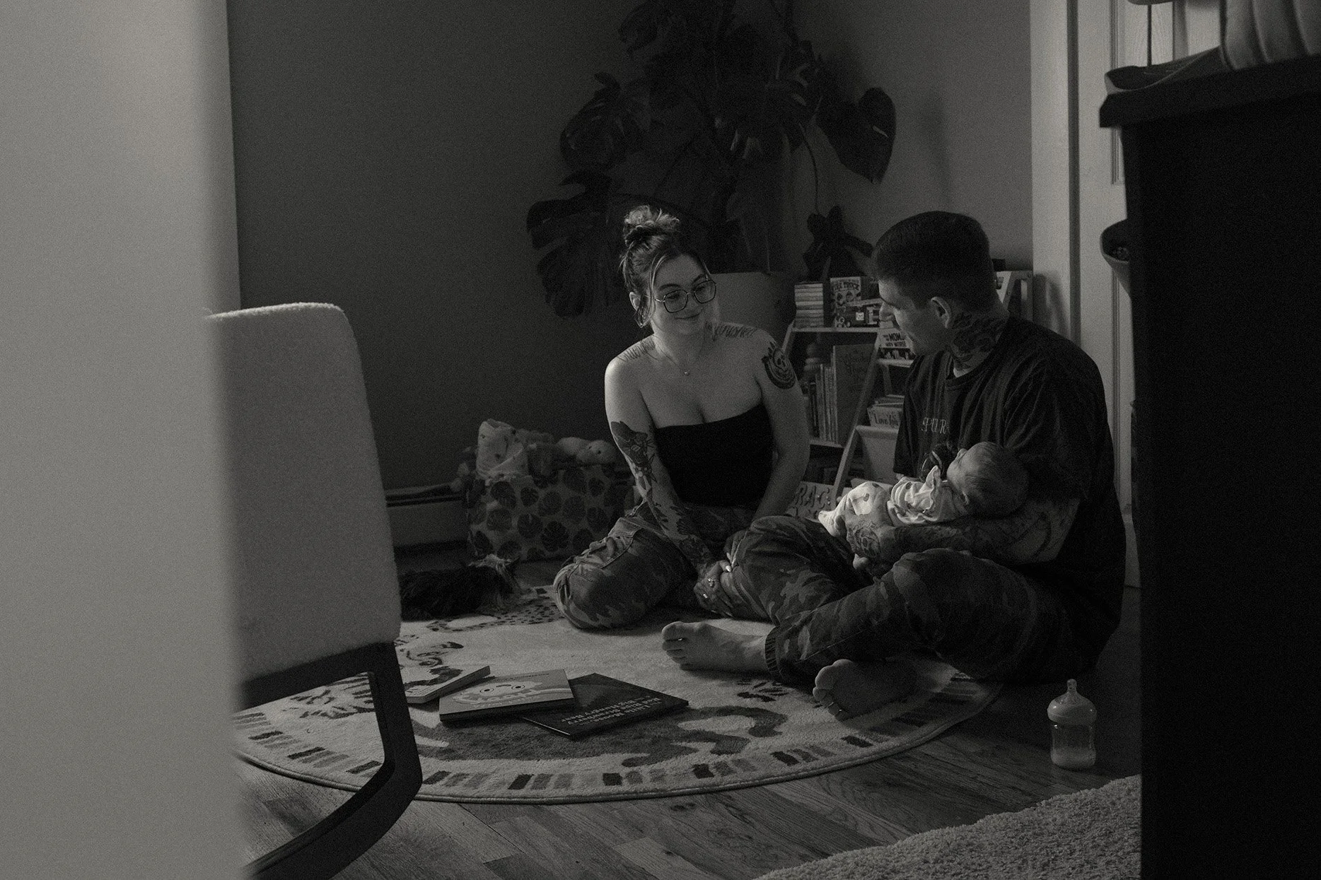 A black and white photo of a woman with glasses and tattoos sitting cross-legged on a rug, holding a small child while talking to a man with tattoos. The man is sitting barefoot on the floor, holding a baby, in a cozy room with a bookshelf and a larg