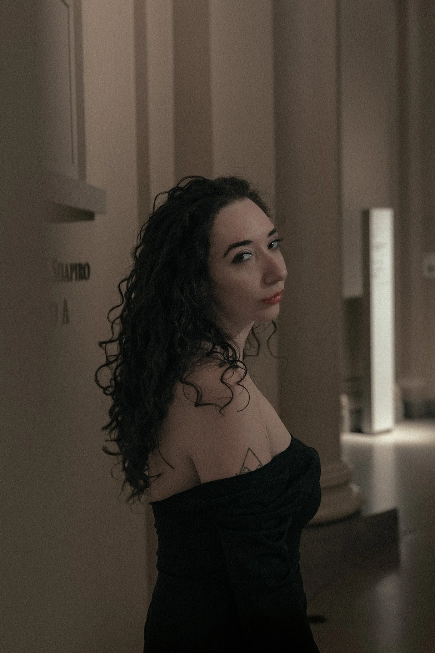 A woman with curly dark hair and light makeup looking over her shoulder in a dimly lit room.