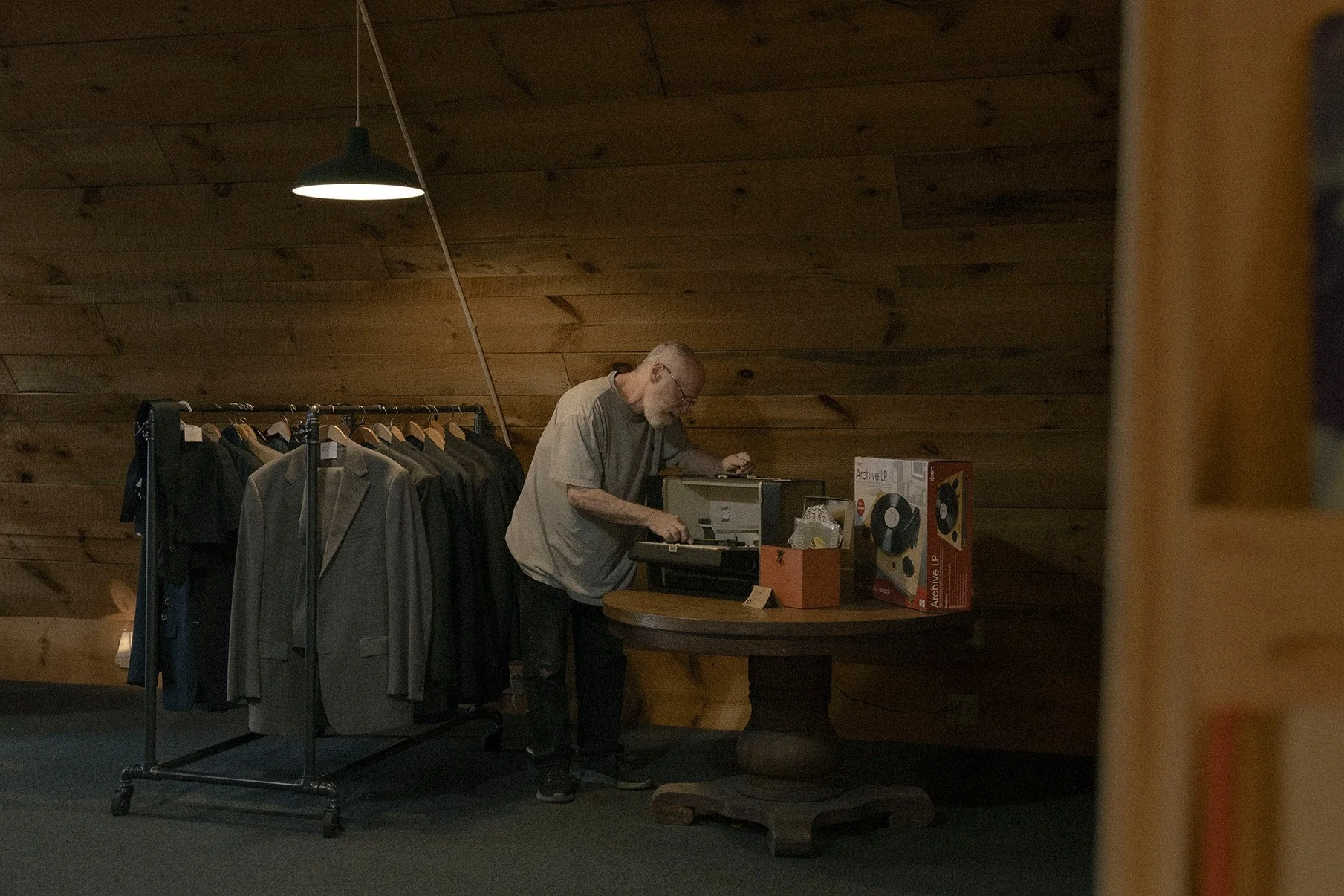 An elderly man with glasses and a beard stands over a wooden table, inspecting a vintage record player in a dimly lit room with wooden walls. To his left, there is a clothing rack with various old fashioned suits and jackets.